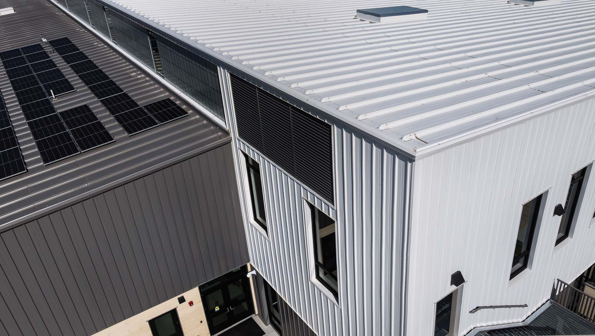 Aerial view of a modern commercial building with white vertical metal panels, standing seam roof and integrated rooftop solar panels showcasing contemporary building envelope design.
