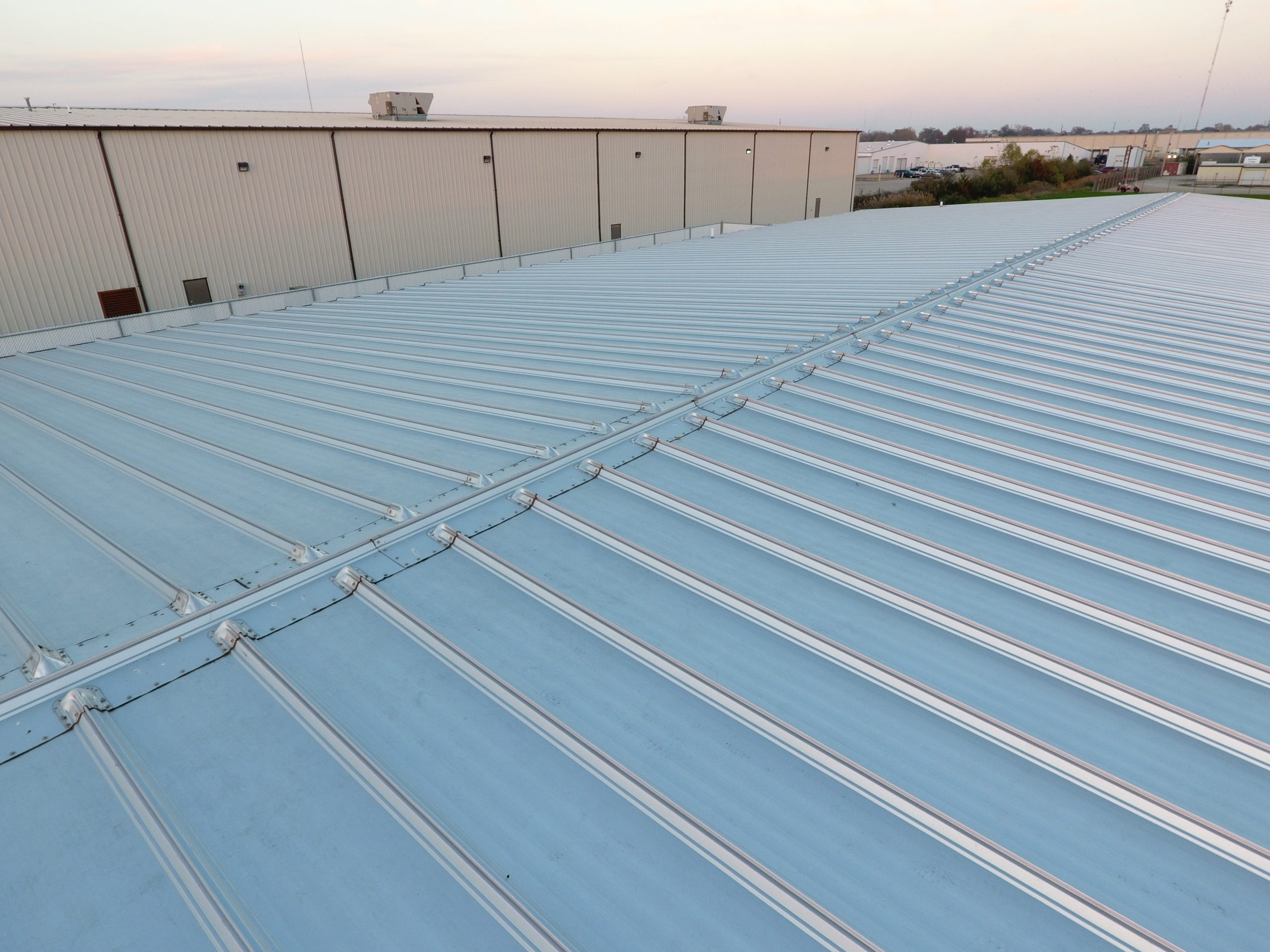 Aerial view of a light blue standing-seam metal roof on a large industrial building, with parallel raised seams running across the surface. Adjacent warehouse structures and a flat industrial landscape are visible in the background under a soft evening sky.