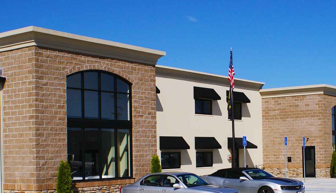 Modern commercial building with tan brick exterior and cream stucco center section, featuring large arched glass entrance, black window awnings, and an American flag on a pole in front. Two cars are parked in the foreground under a clear blue sky.