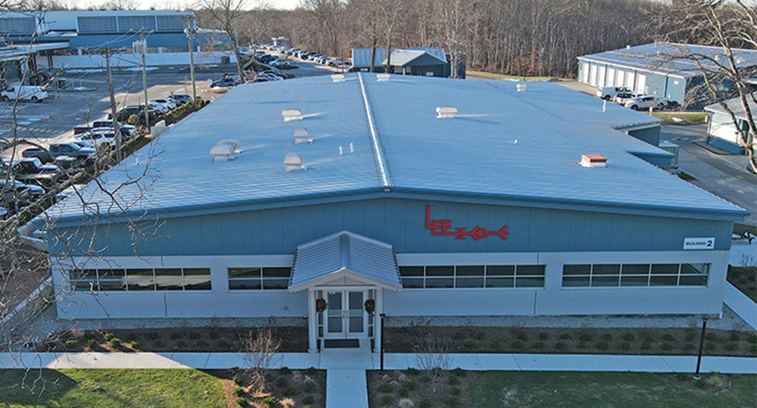 Aerial front view of a blue metal industrial building with a standing-seam roof and central entrance canopy. The facade features long horizontal windows and a red “Lee” logo, with parking lots, neighboring industrial buildings, and leafless trees surrounding the site.