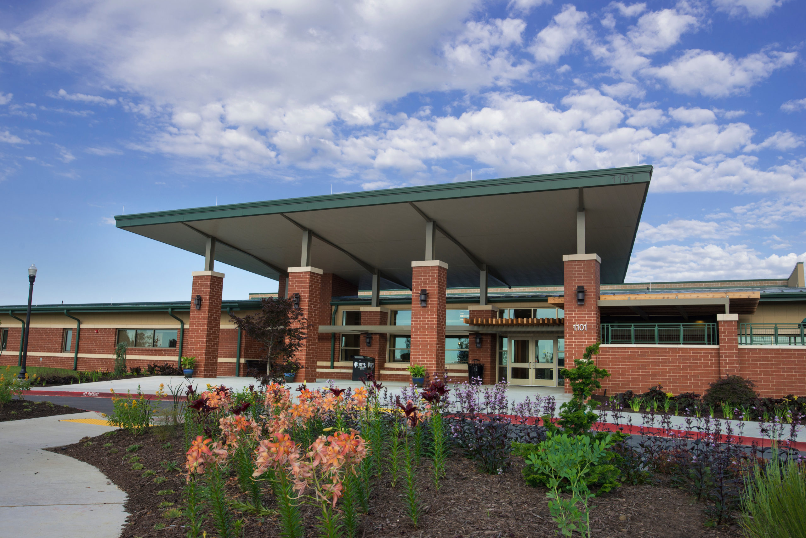 Exterior of a modern brick community or institutional building with large covered entrance canopy, landscaped garden beds, and glass entry doors under a partly cloudy sky.