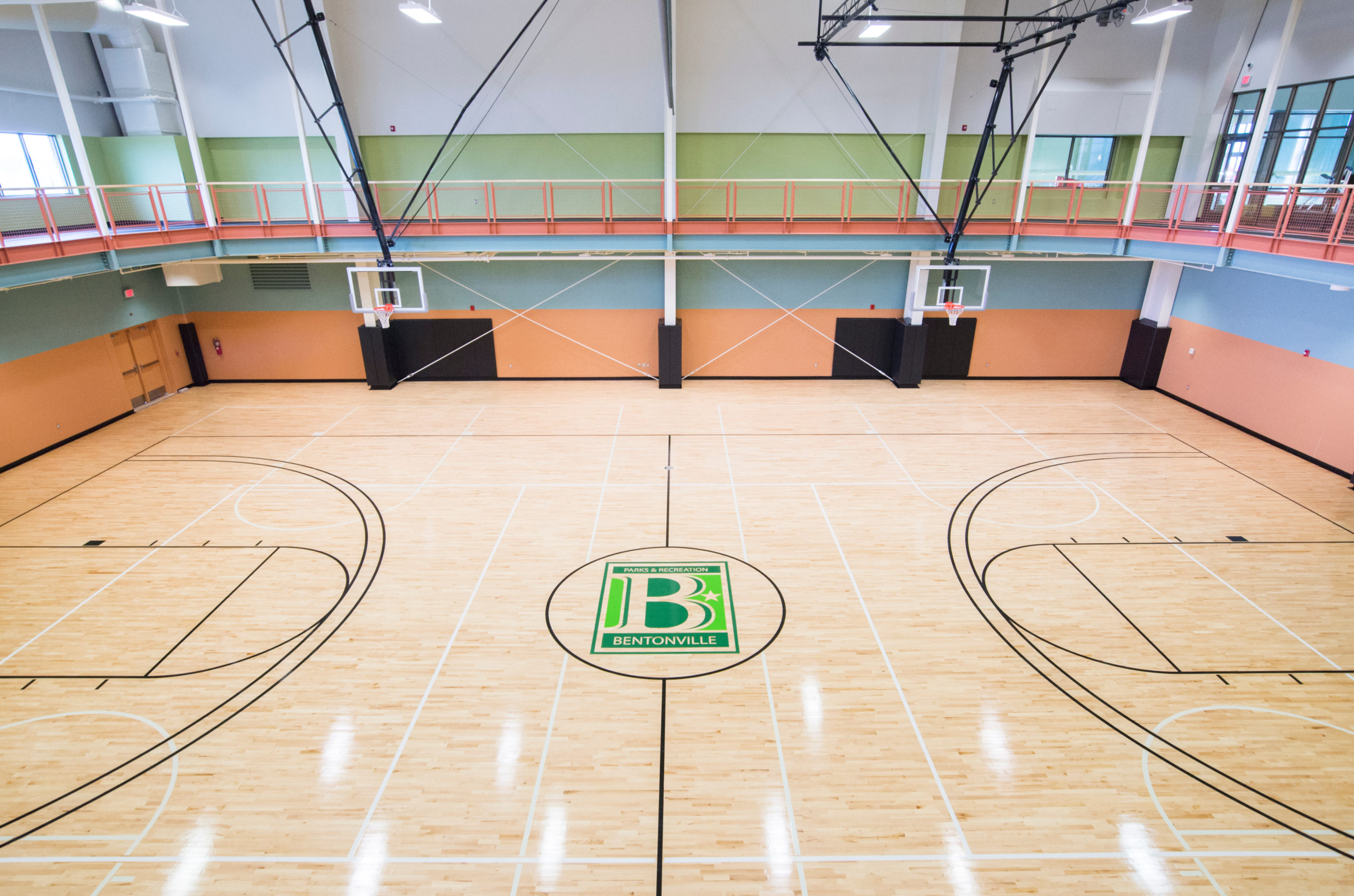 Interior view of a modern indoor basketball court with wood flooring, multiple hoops, elevated walking track, and Bentonville Parks & Recreation logo at center court.