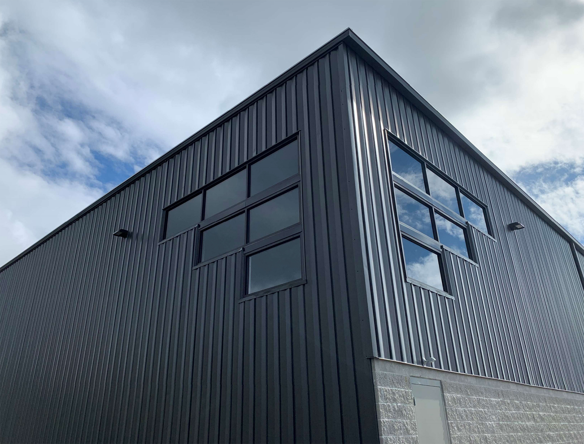 Exterior corner view of a modern metal building with dark vertical panel siding, large rectangular windows, and a concrete block base under a cloudy sky.