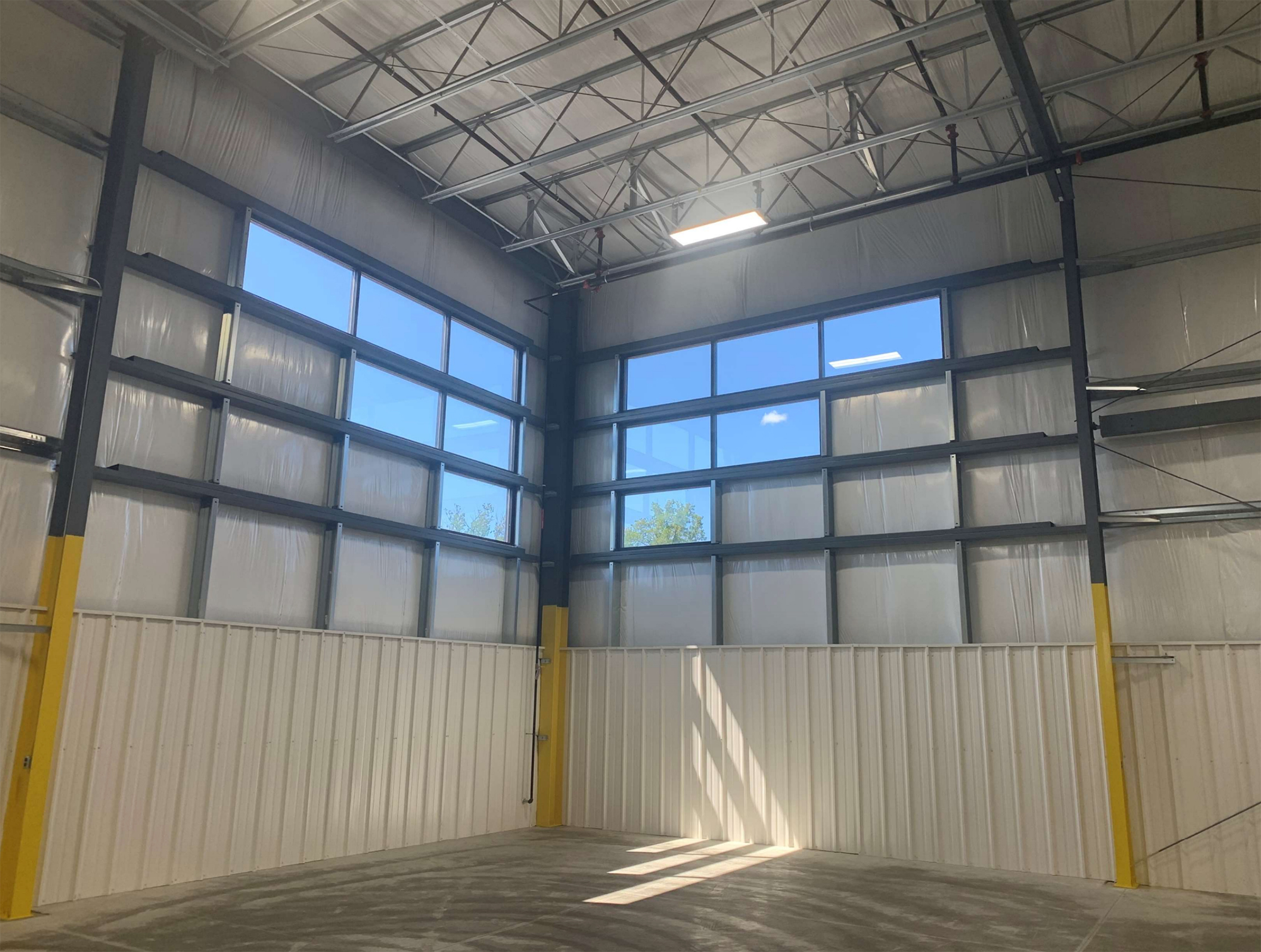 Interior corner of a metal building with exposed steel framing, high clerestory windows, insulated wall panels, and polished concrete floor lit by natural sunlight.
