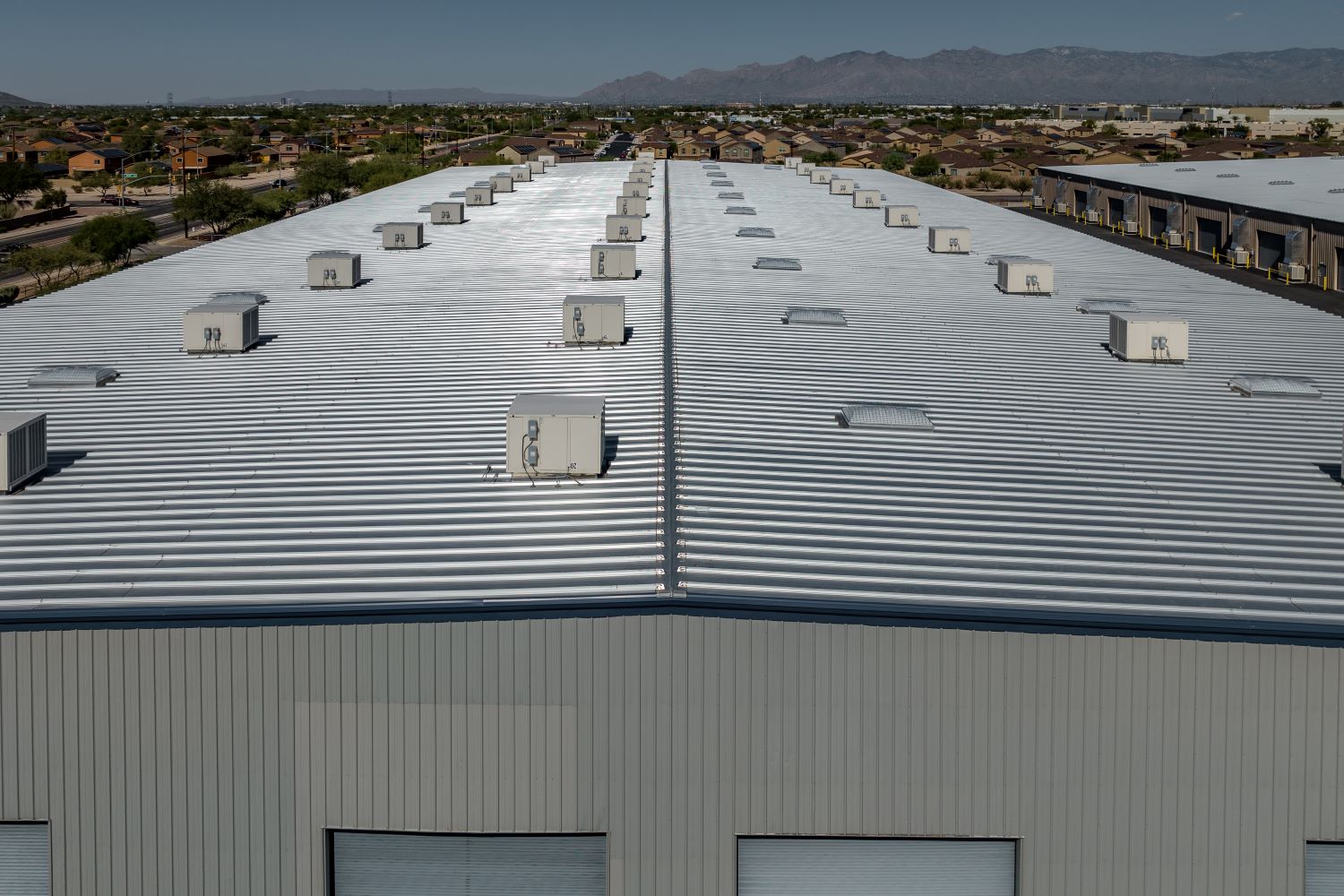 Aerial view of a large metal-roof industrial building with multiple rooftop HVAC units and skylights arranged in rows, surrounded by a suburban neighborhood and distant mountains under a clear sky.