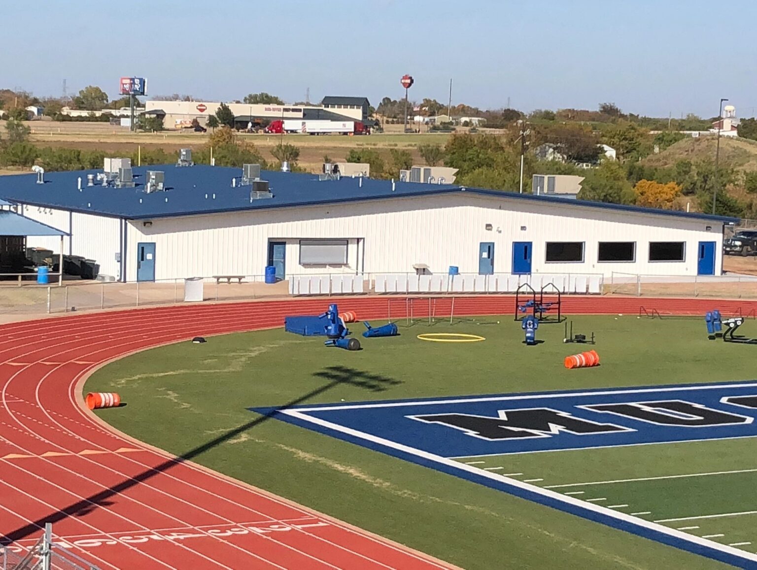 Athletic field with a red running track surrounding green turf marked with large blue lettering, training equipment scattered on the field, and a long metal-sided school or sports facility building in the background under a clear sky.