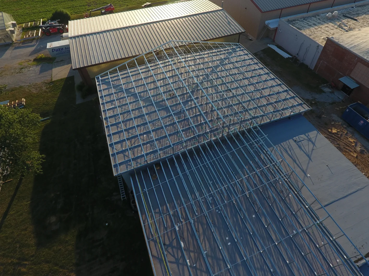 Aerial view of a steel-frame building under construction, showing exposed metal roof trusses and structural grid extending from an existing metal-roof facility, with surrounding industrial buildings and equipment nearby.