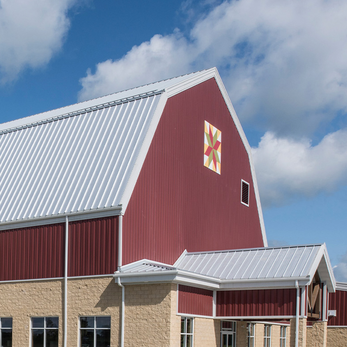 Red and tan agricultural facility with white metal roof, central silo feature and expansive glass entry beneath a bright sky with scattered clouds.