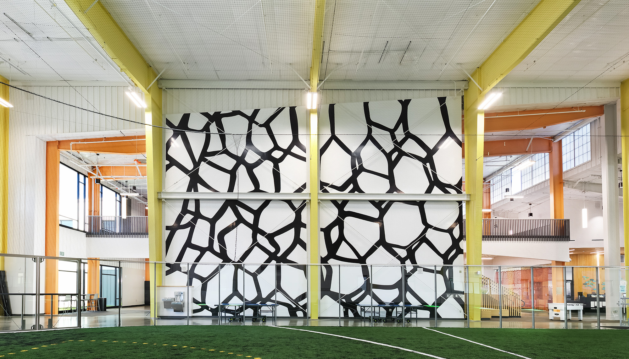 Indoor athletic and recreation space with bright yellow structural framing, open mezzanine levels and large graphic feature wall surrounding a turf activity area.