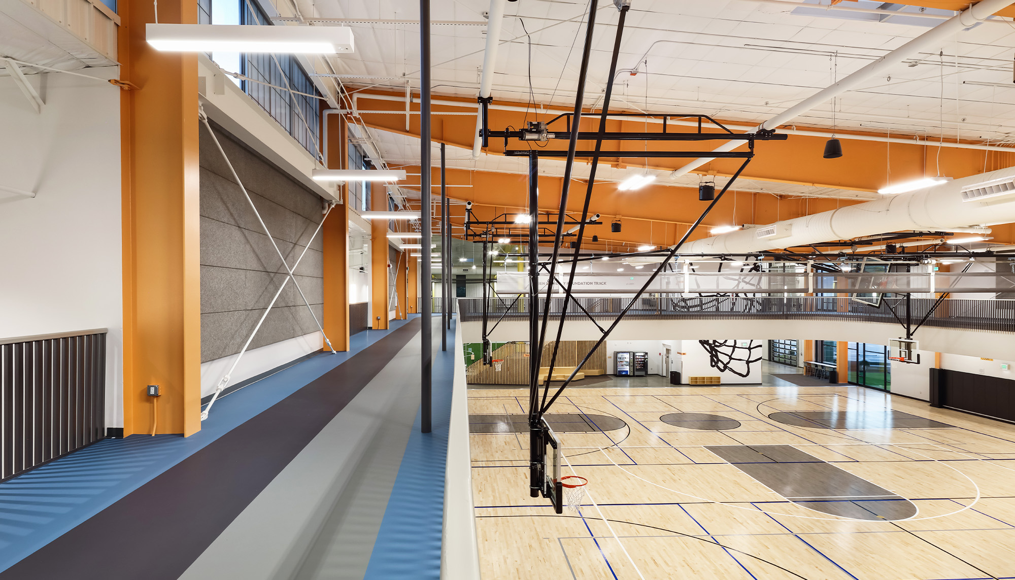 Contemporary indoor recreation facility with elevated walking track, warm orange structural framing and open multi-court athletic space below.