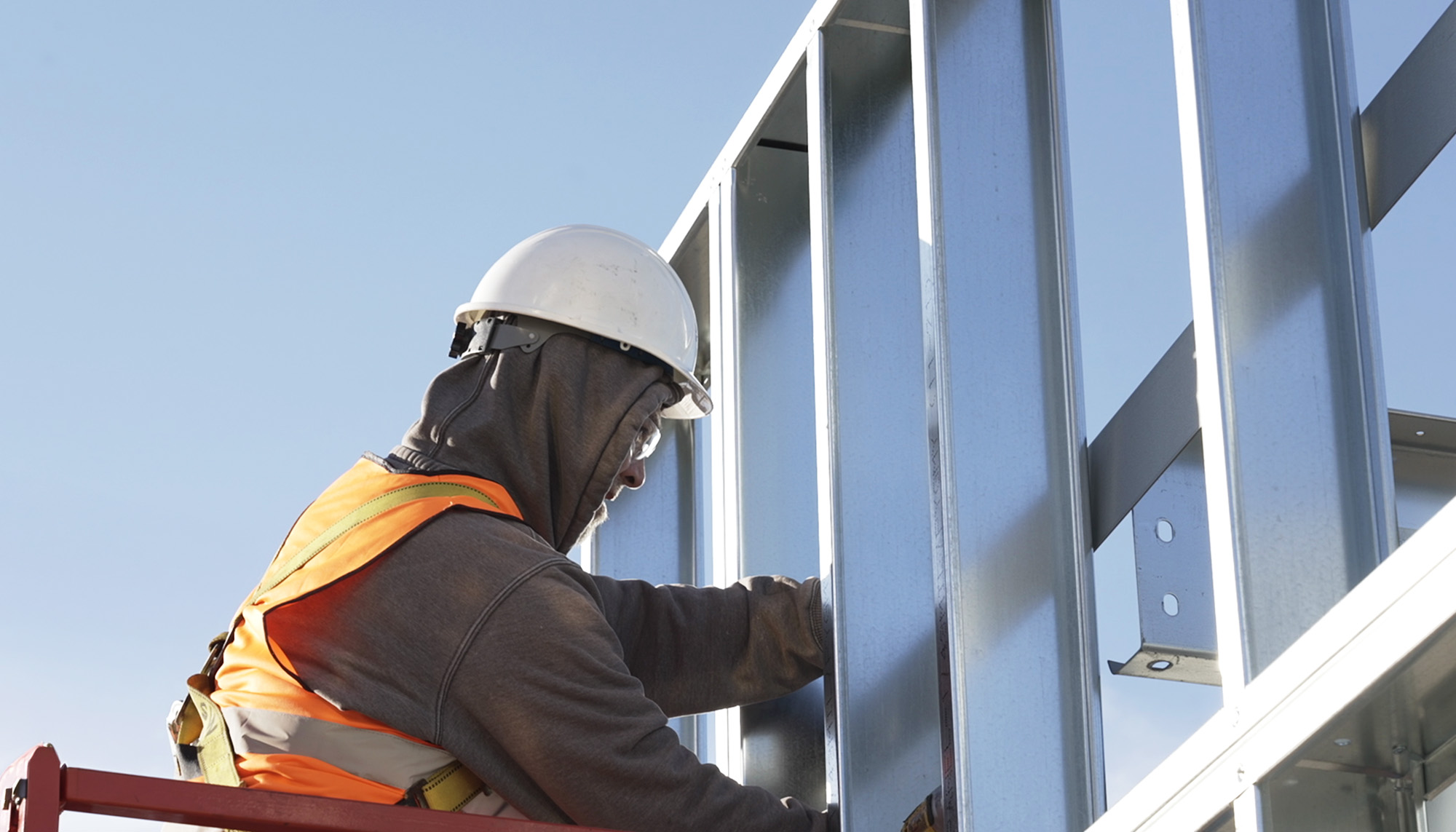 Construction worker in a safety vest and hard hat installing metal wall framing on a building exterior.