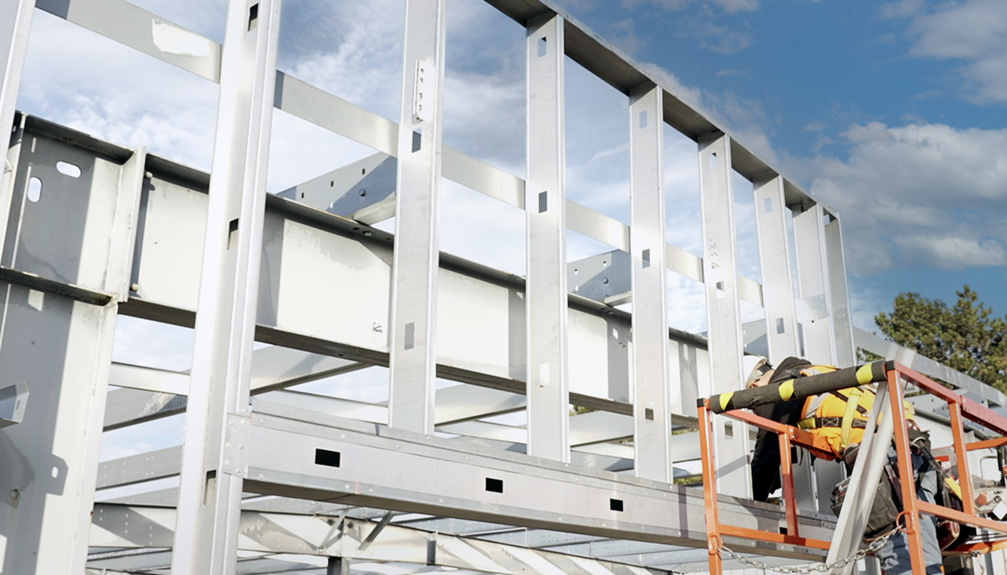 Worker on a lift installing structural steel framing for a metal building under a blue sky.