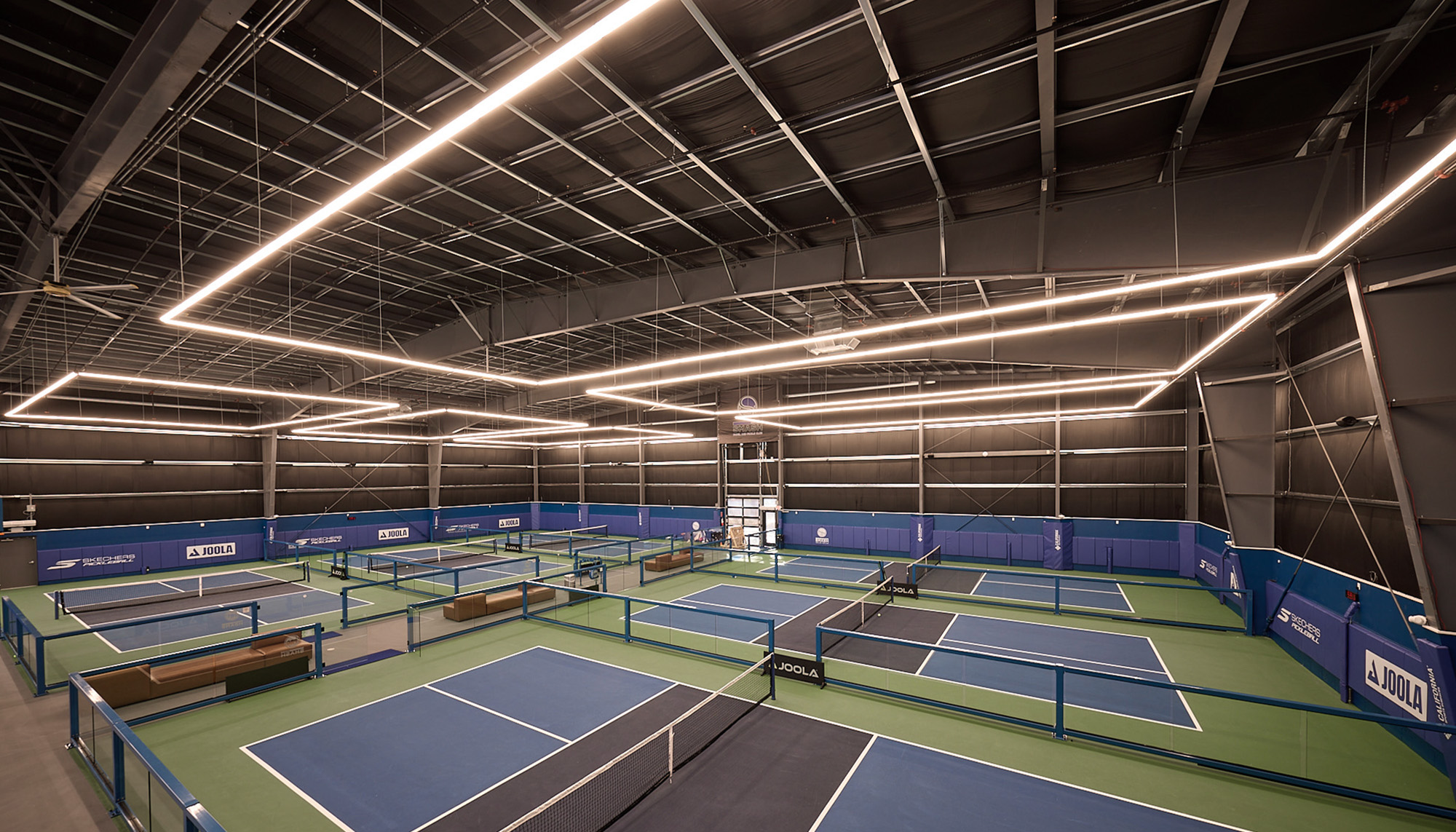 Indoor pickleball facility with multiple blue-and-green courts, exposed steel ceiling structure and modern geometric lighting throughout.