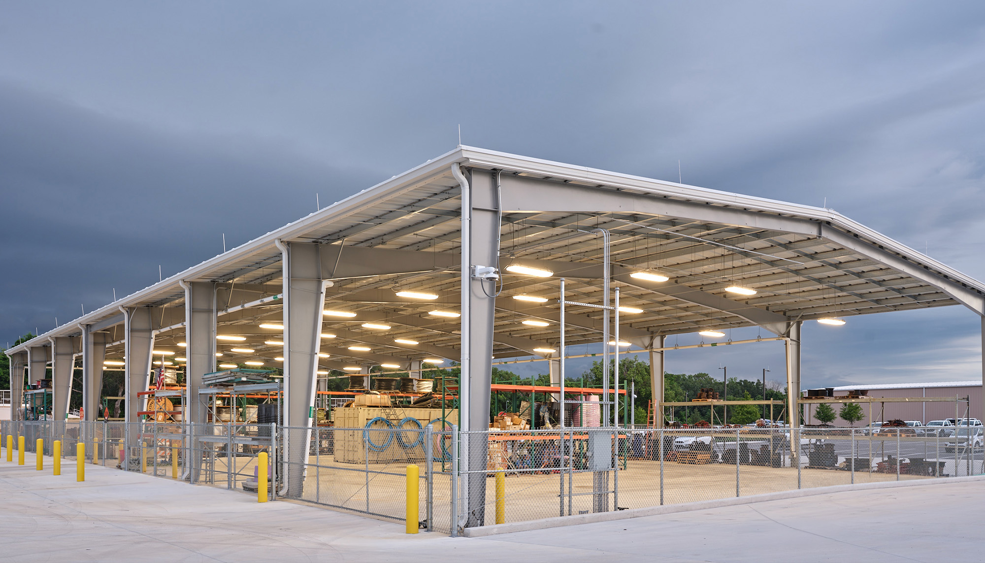 Open-air steel canopy structure with illuminated covered workspace, exposed framing and integrated utility access beneath a dramatic evening sky.