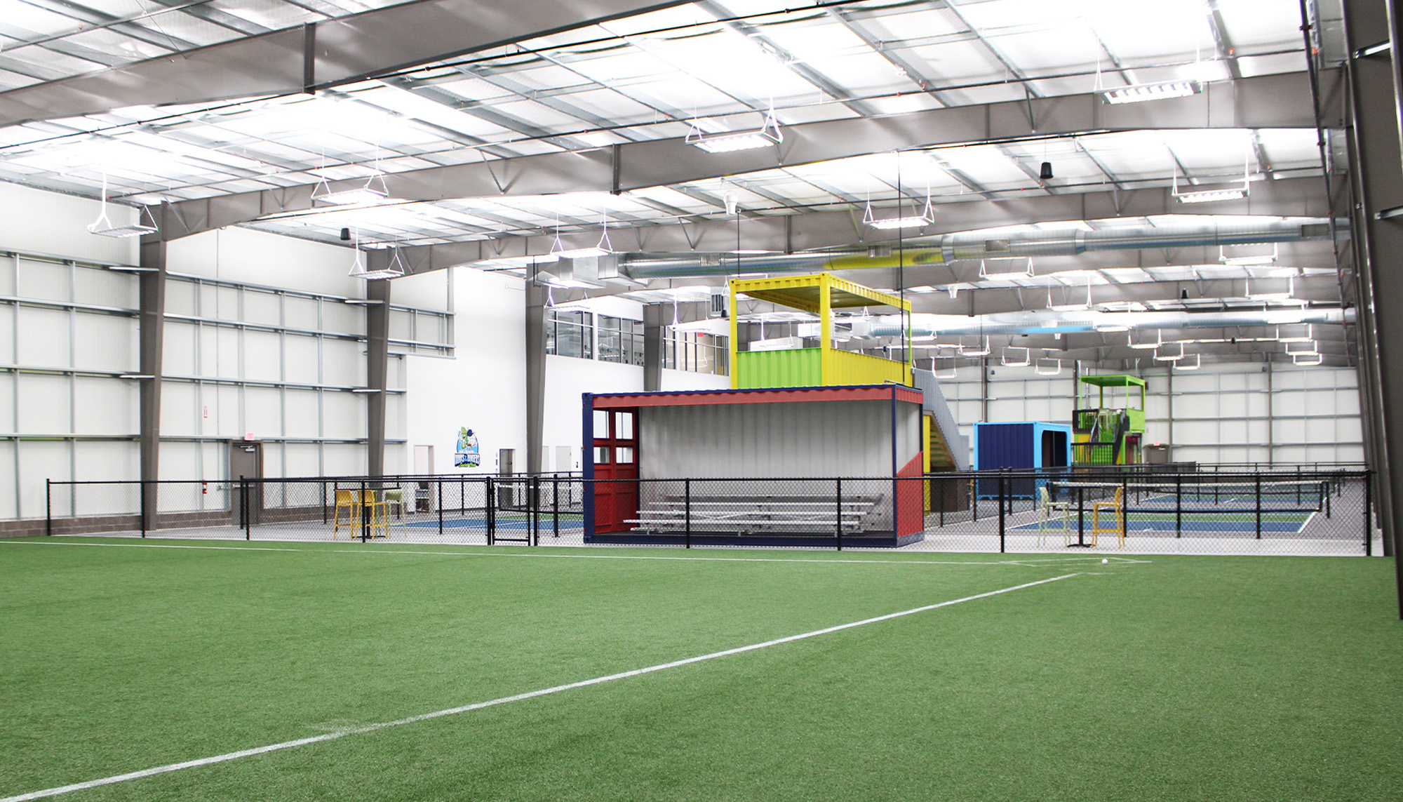 Indoor sports facility with green artificial turf, white metal walls and ceiling, black netting, and a red-and-gray enclosed structure beneath bright overhead lighting.