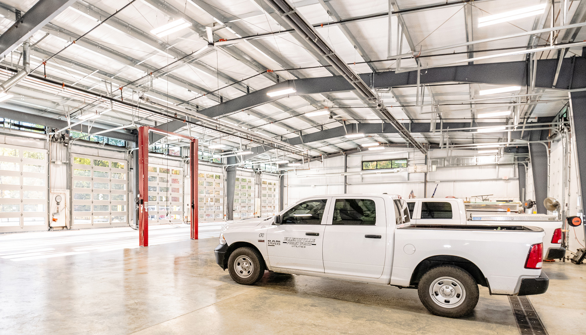 Bright industrial garage interior with white metal walls and ceiling, polished concrete floors, red vehicle lifts, and a white service pickup truck parked inside.