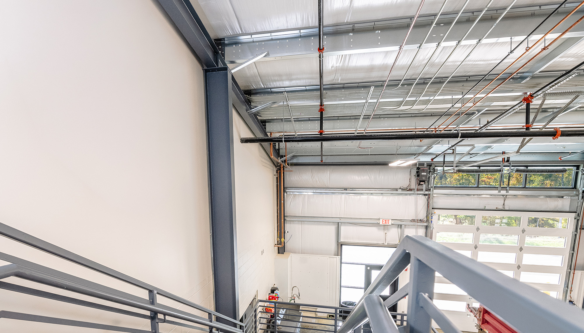 Interior stairwell in a steel-framed industrial building with gray railings, white insulated wall panels, exposed piping and conduit, and natural light from upper windows.