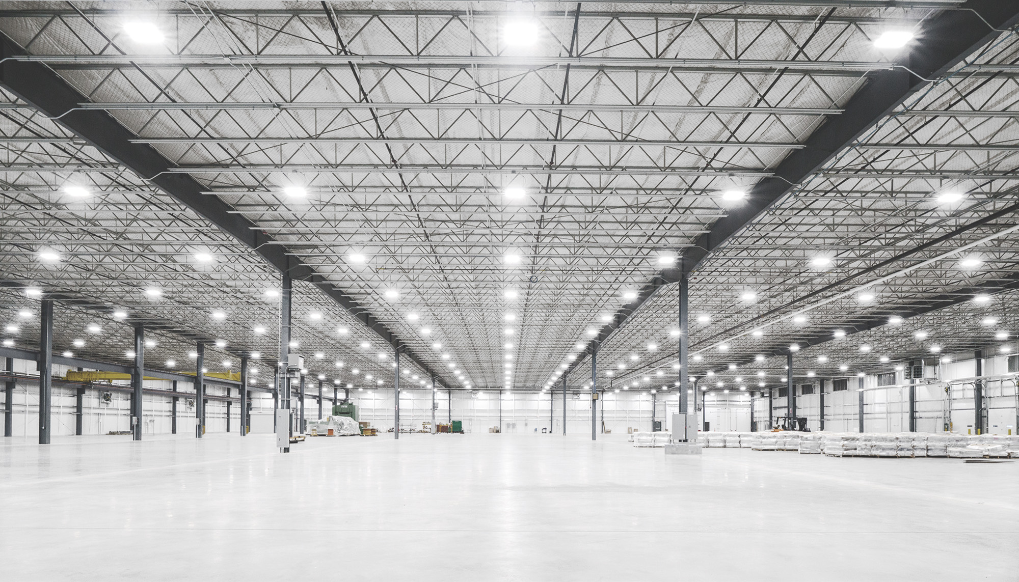 Expansive warehouse interior with white concrete floors, black steel framing, exposed metal trusses, and rows of bright overhead lights extending across the ceiling.