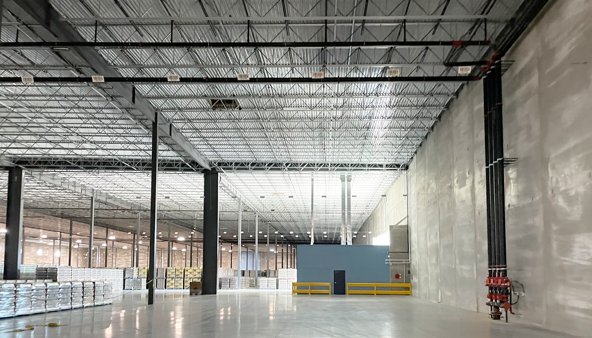 Large industrial warehouse interior with polished concrete floors, black steel columns, white insulated wall panels, exposed metal roof decking, and bright overhead lighting.
