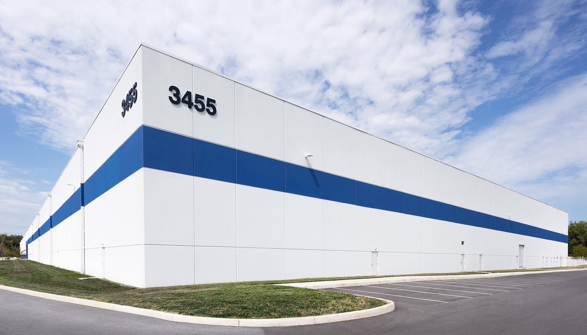 White industrial warehouse building with a bold blue horizontal stripe, black building numbers, and a clean exterior under a partly cloudy sky.