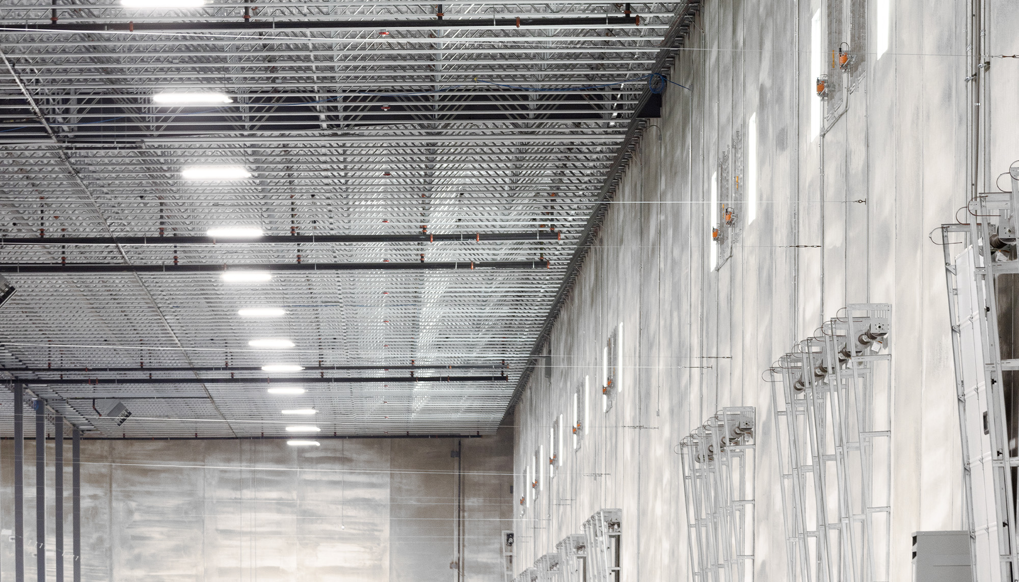 Industrial warehouse interior with tall gray concrete wall panels, exposed steel ceiling structure, suspended lighting, and polished concrete floors.