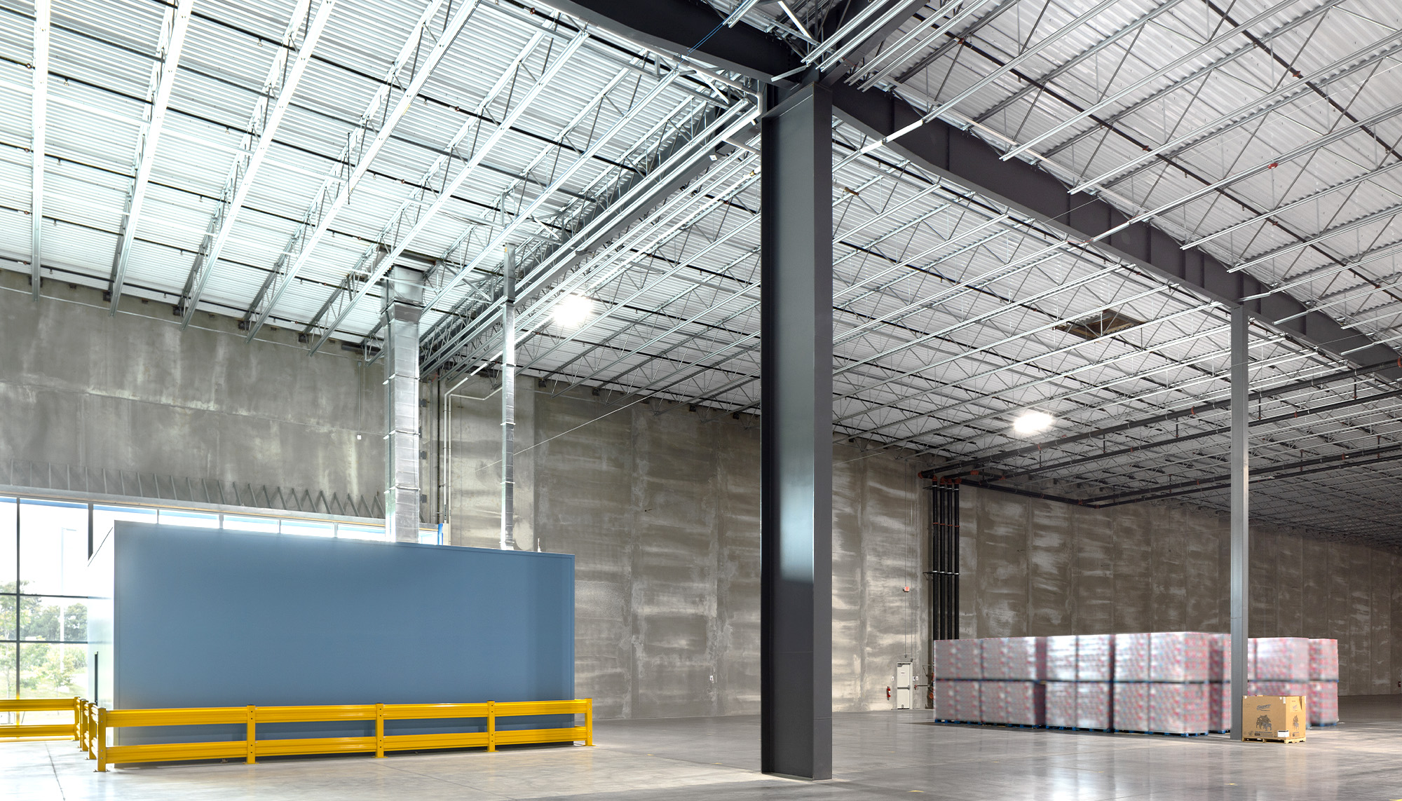 Spacious warehouse interior with gray concrete walls, black steel columns, white insulated ceiling panels, yellow safety railings, and stacked palletized materials.