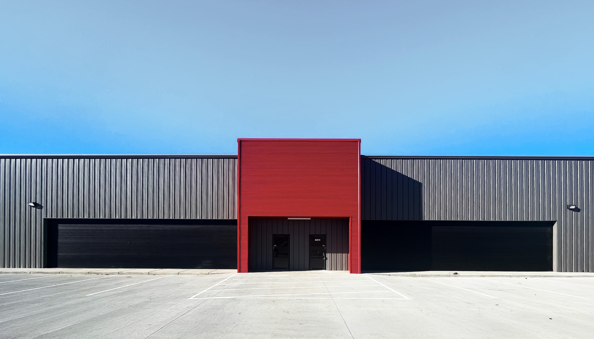 Front view of a black metal building with a bold red central entry, symmetrical garage bays and wide concrete apron.