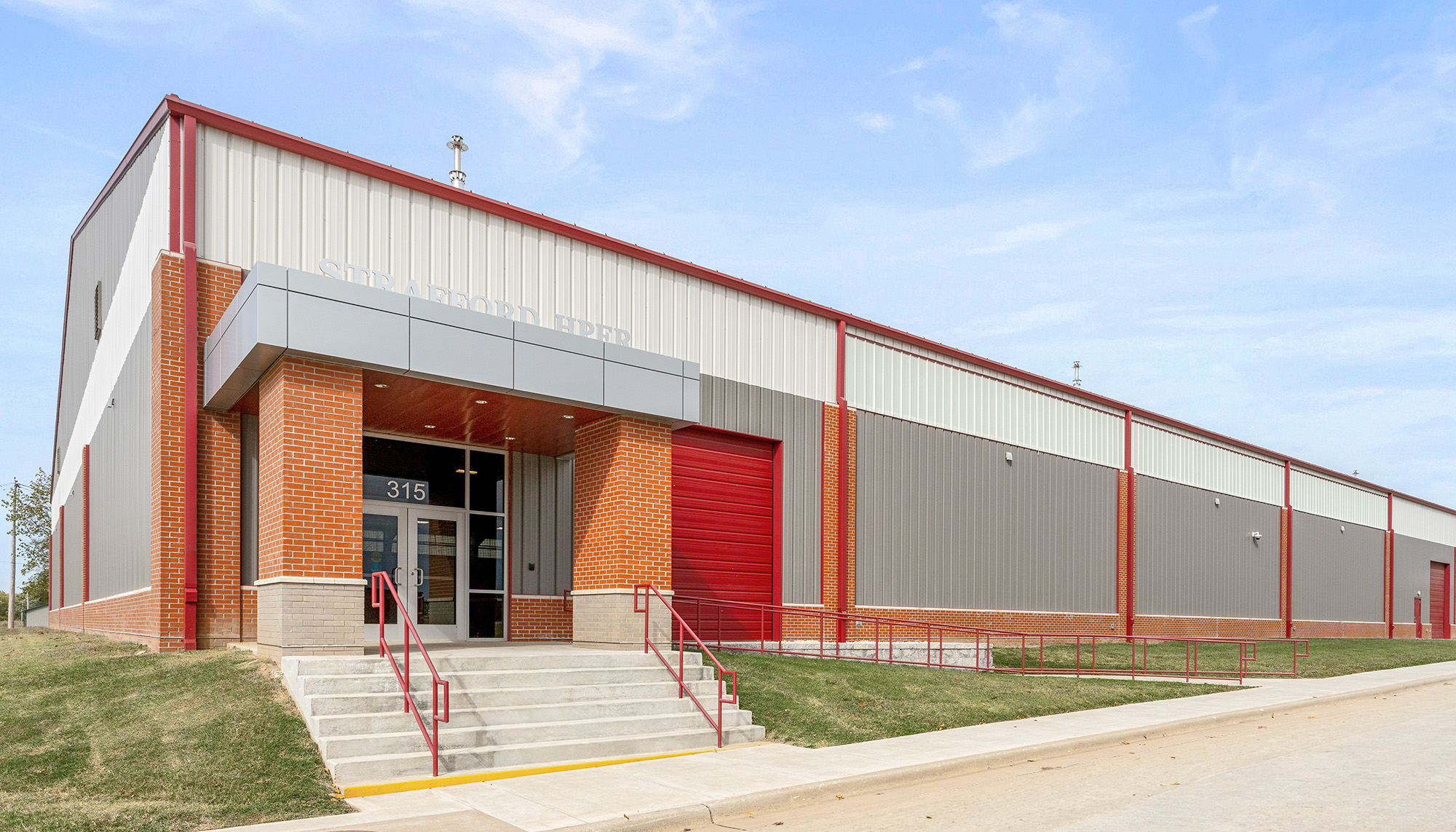Large metal building with brick office entry, red overhead door and extended sidewall panels along a sloped site.