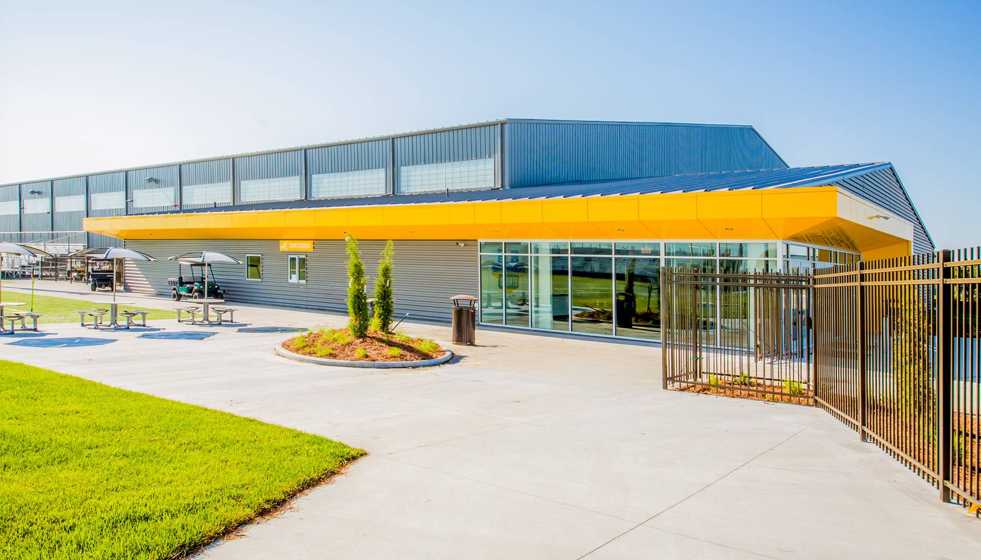 Modern metal building with blue upper panels, yellow canopy and glass façade opening to a landscaped outdoor seating area.