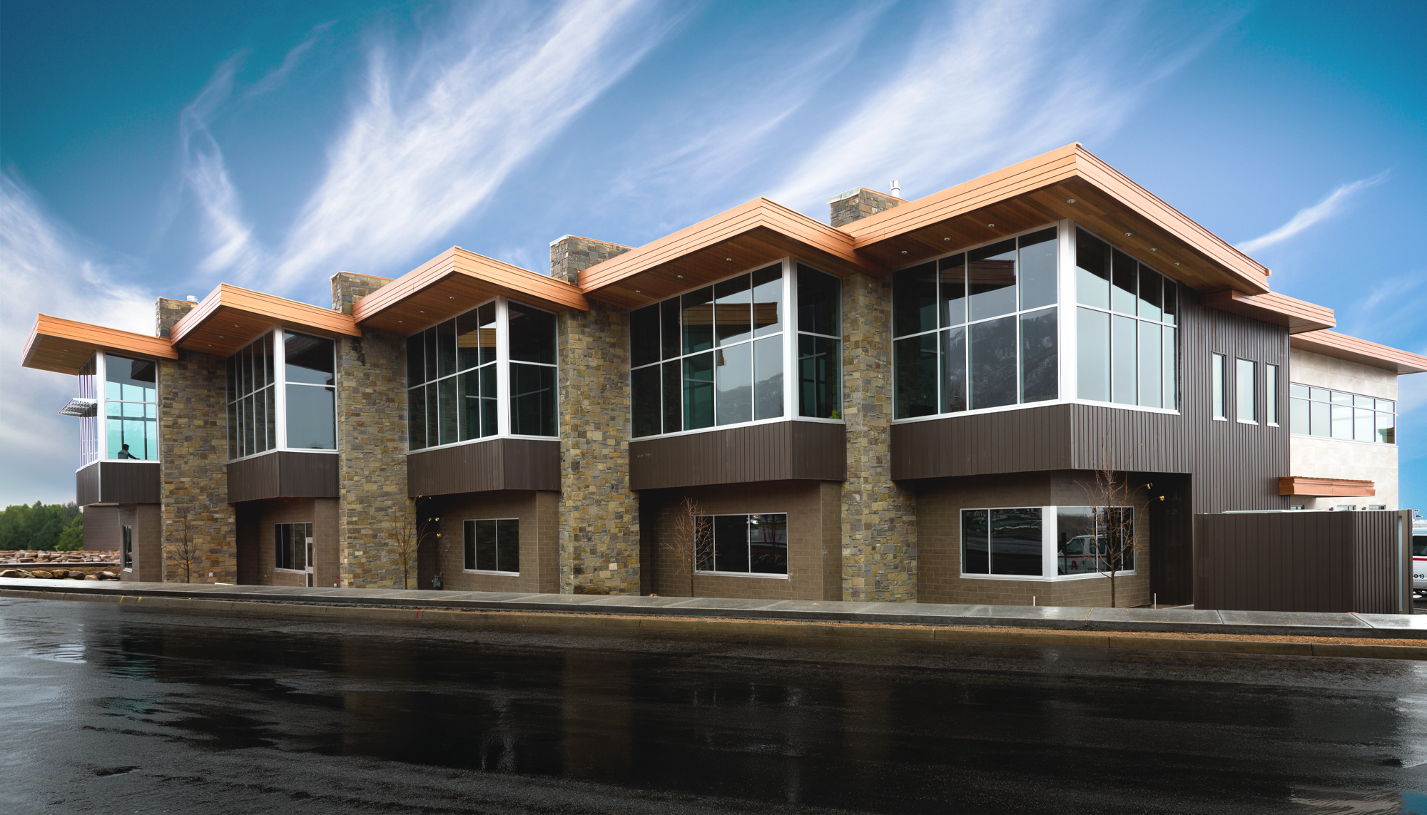 Two-story commercial building with stone and metal façade, large corner windows and layered rooflines along a wet street frontage.