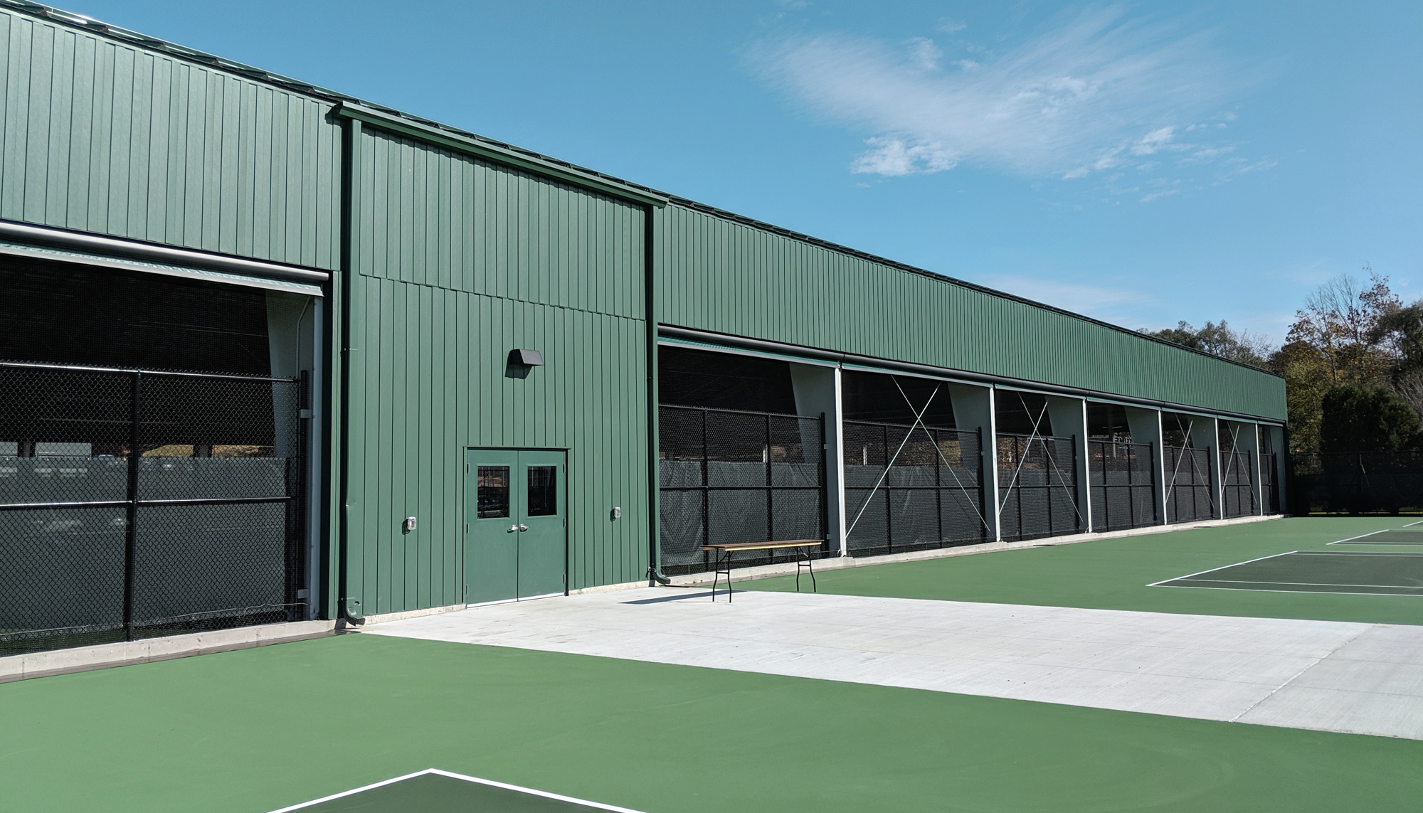 Long green metal building with open bay sections, adjacent to outdoor sport courts and paved walkways.