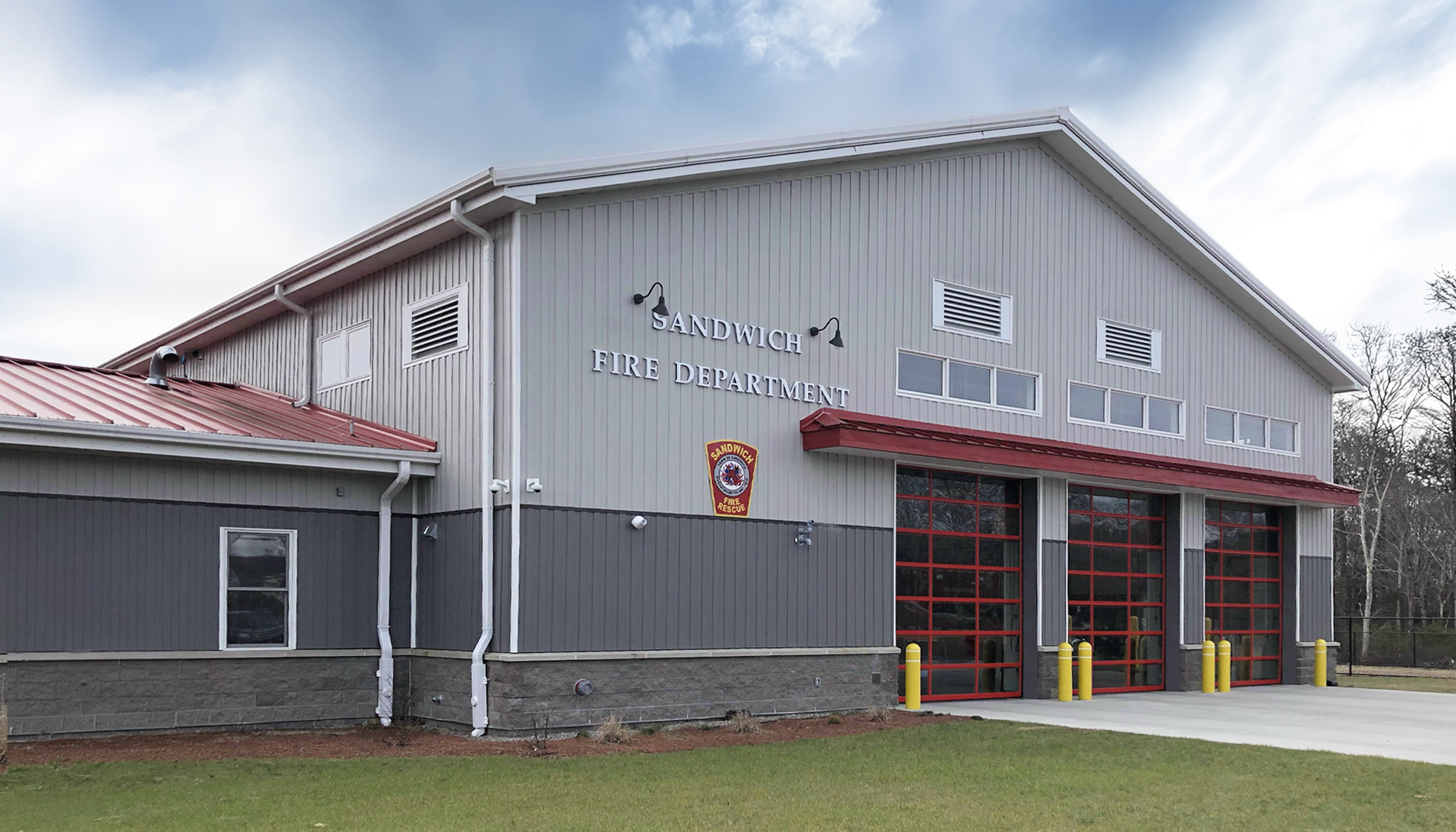 Light gray metal fire station building with red bay doors, stone base accents and a small front lawn.