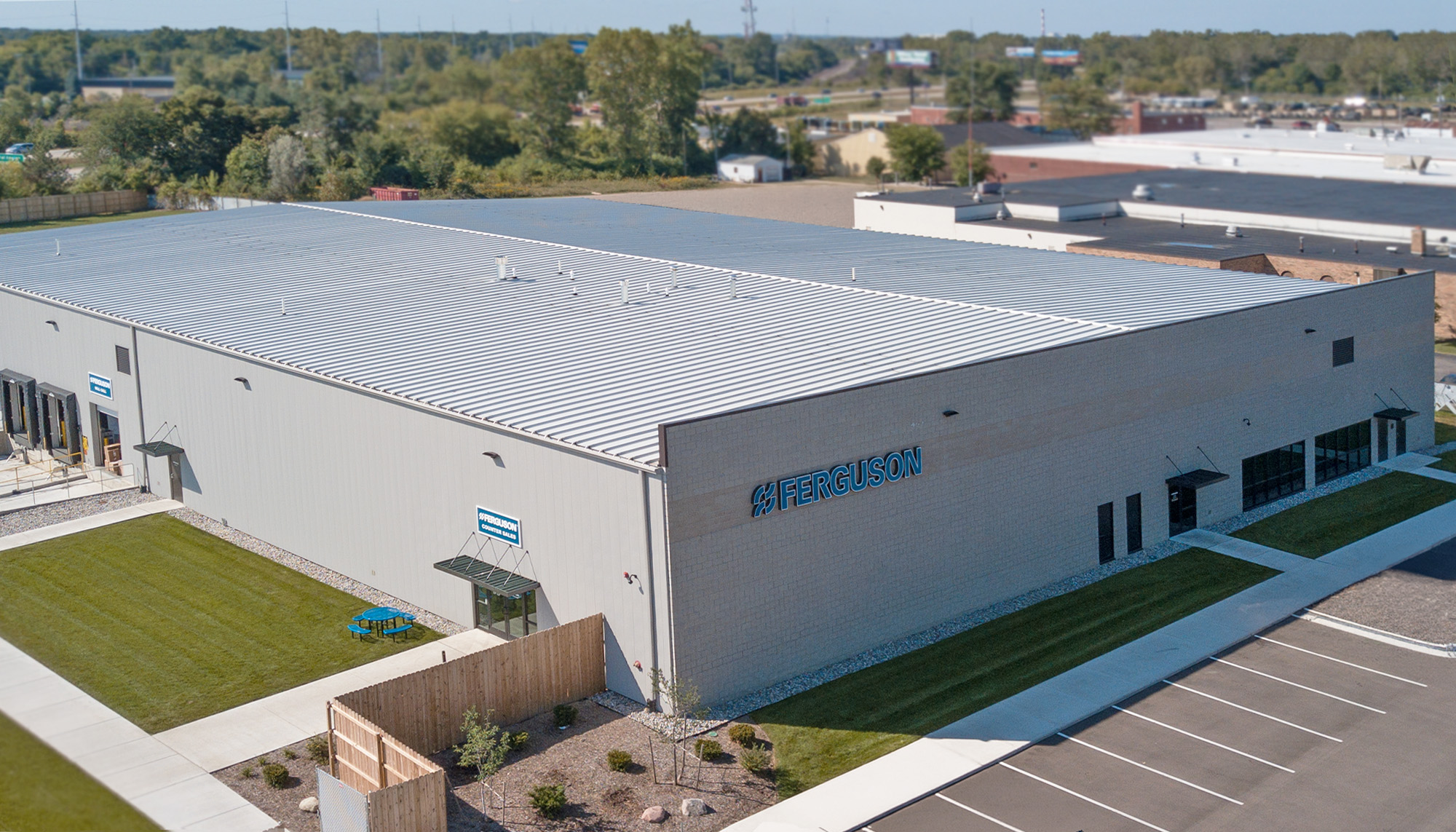 Aerial view of a large light-gray metal industrial building with standing seam roof, adjacent parking and landscaped perimeter.