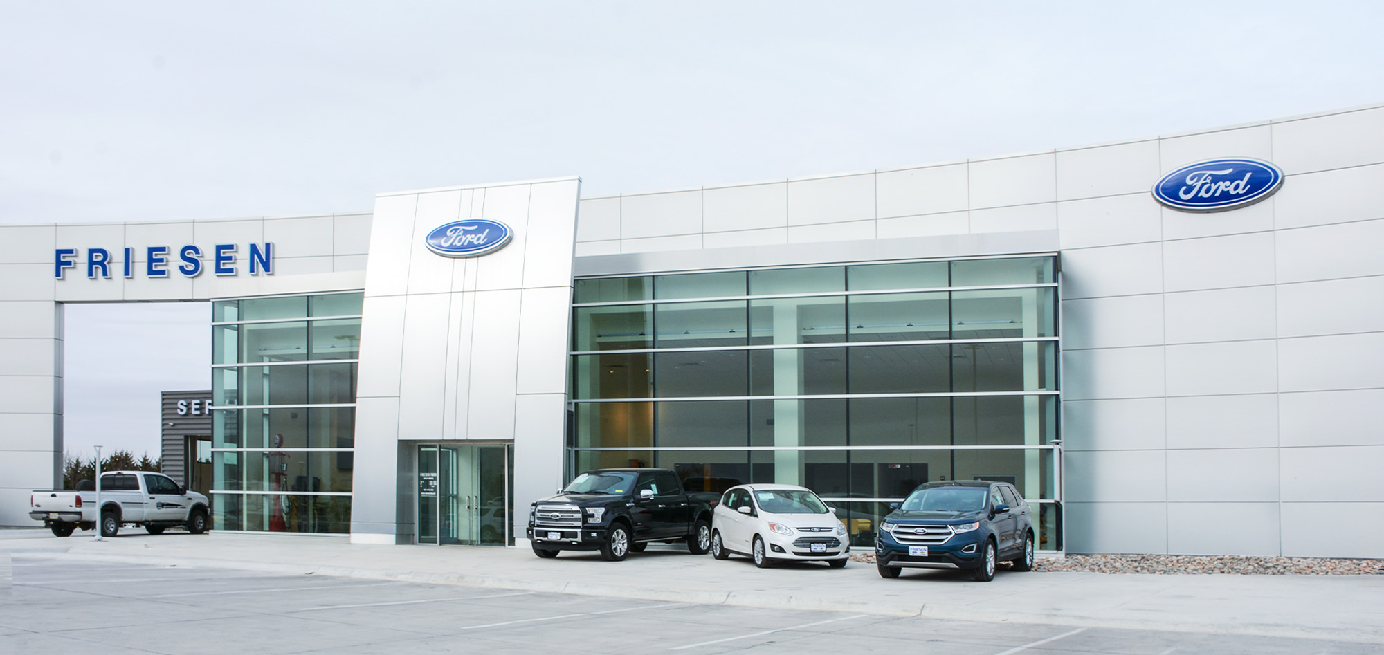 Modern white automotive dealership with expansive glass showroom, clean architectural lines and branded entry tower set against an overcast sky.