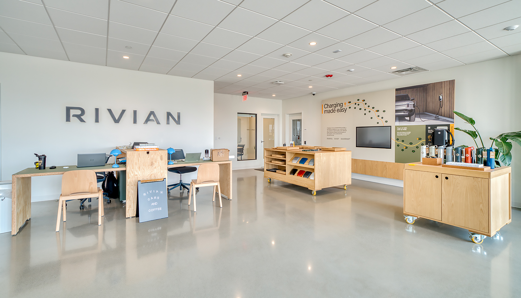 Bright RIVIAN showroom with white walls, polished gray floors, light wood furniture, product displays and green plants.