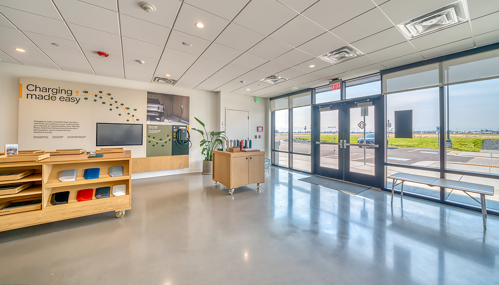 Bright RIVIAN showroom with polished gray floors, light wood displays, green plants, blue-framed glass doors and large windows overlooking the exterior.