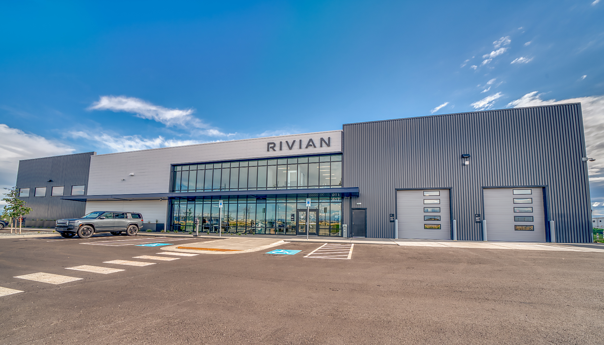Modern gray RIVIAN building with a glass-fronted entrance, two garage doors and marked parking spaces under a bright blue sky.
