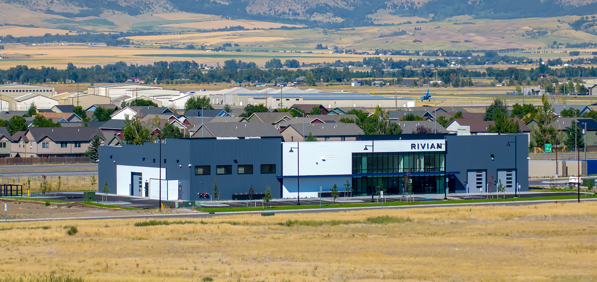 Modern gray and white RIVIAN industrial building with large windows, surrounded by golden fields, nearby homes and rolling hills.