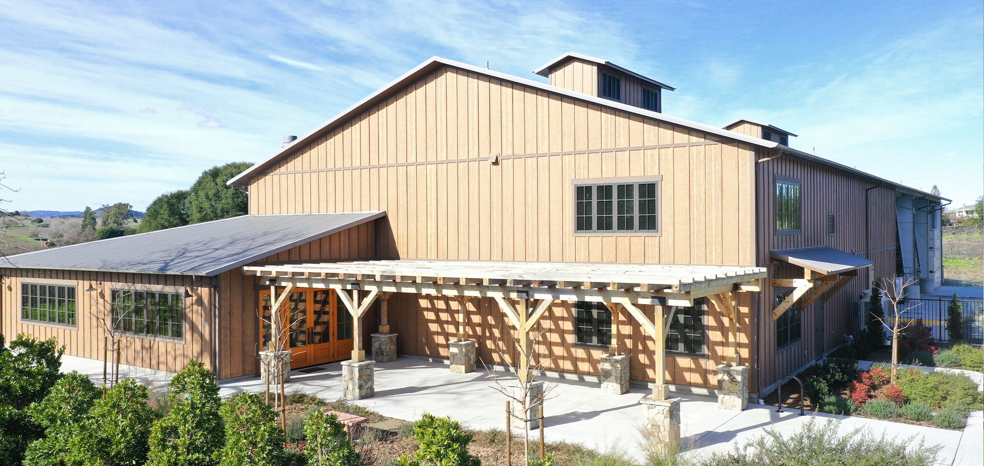 Rustic tan agricultural and event facility with timber-framed covered porch, metal rooflines and landscaped outdoor gathering spaces set against a scenic rural backdrop.