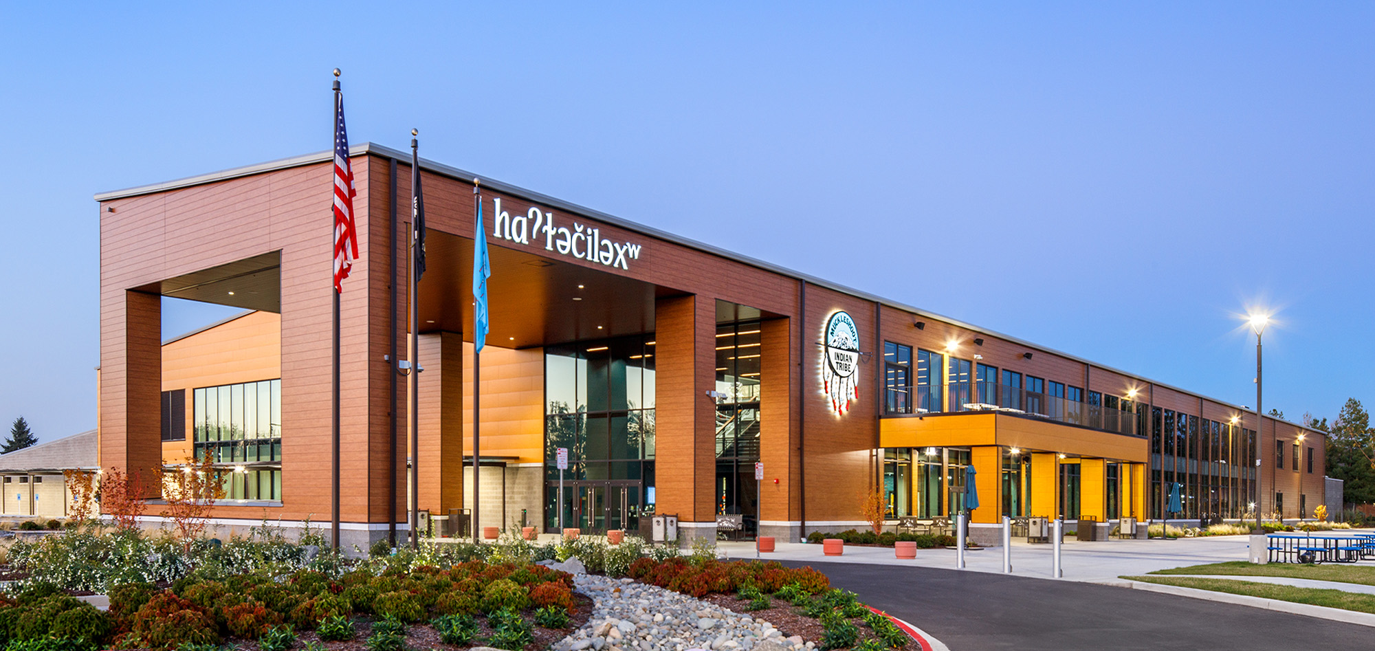 Contemporary civic building with warm wood-tone facade, expansive glass entry and landscaped frontage illuminated against a clear evening sky.
