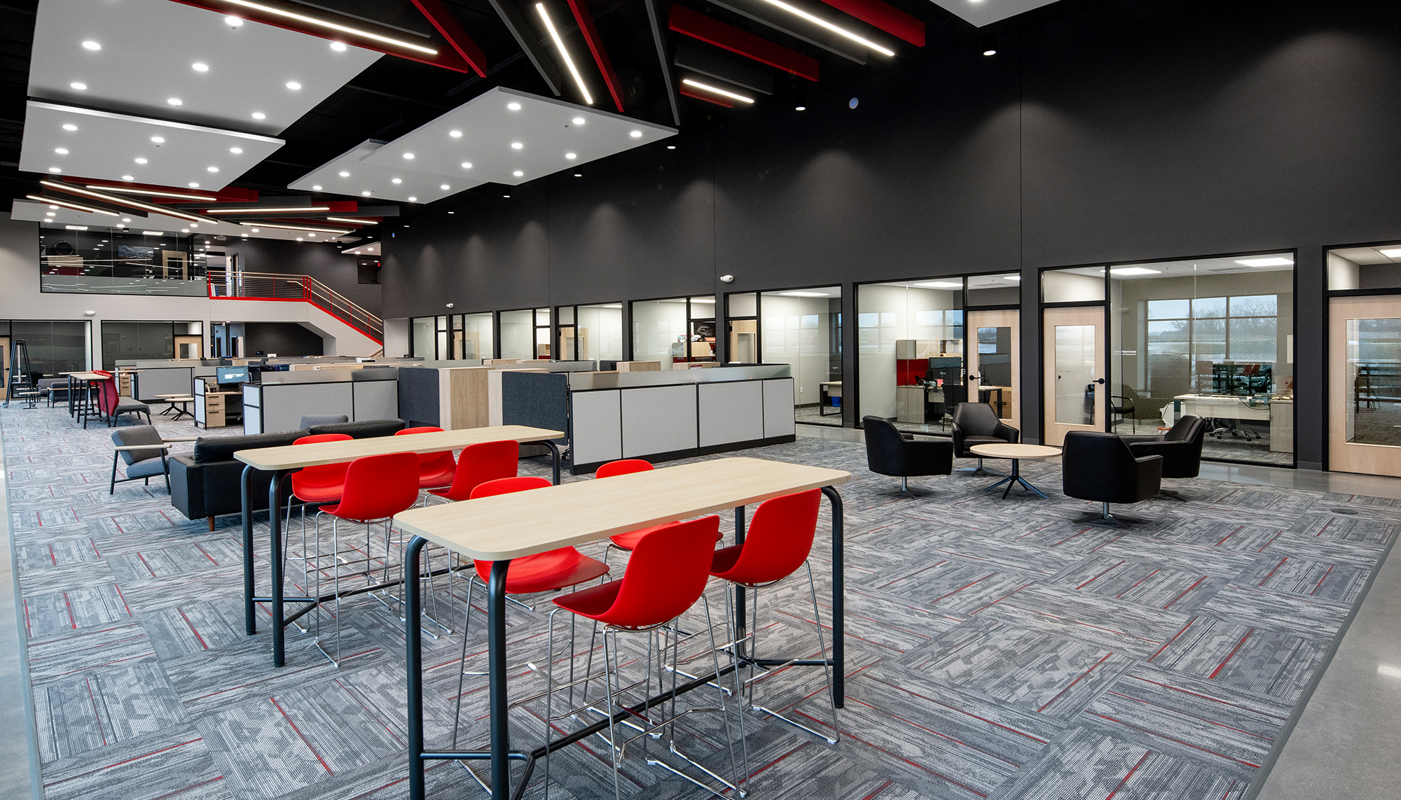 Modern open office with gray patterned carpet, black walls, red chairs and accents, shared workstations and geometric ceiling lights.