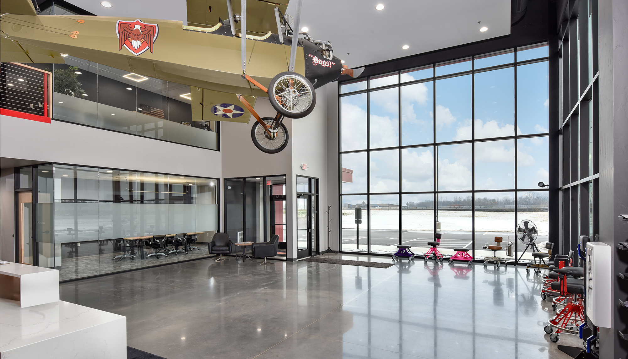 Modern industrial lobby with polished gray floors, black steel framing, red accents, floor-to-ceiling windows and a suspended aircraft display overhead.