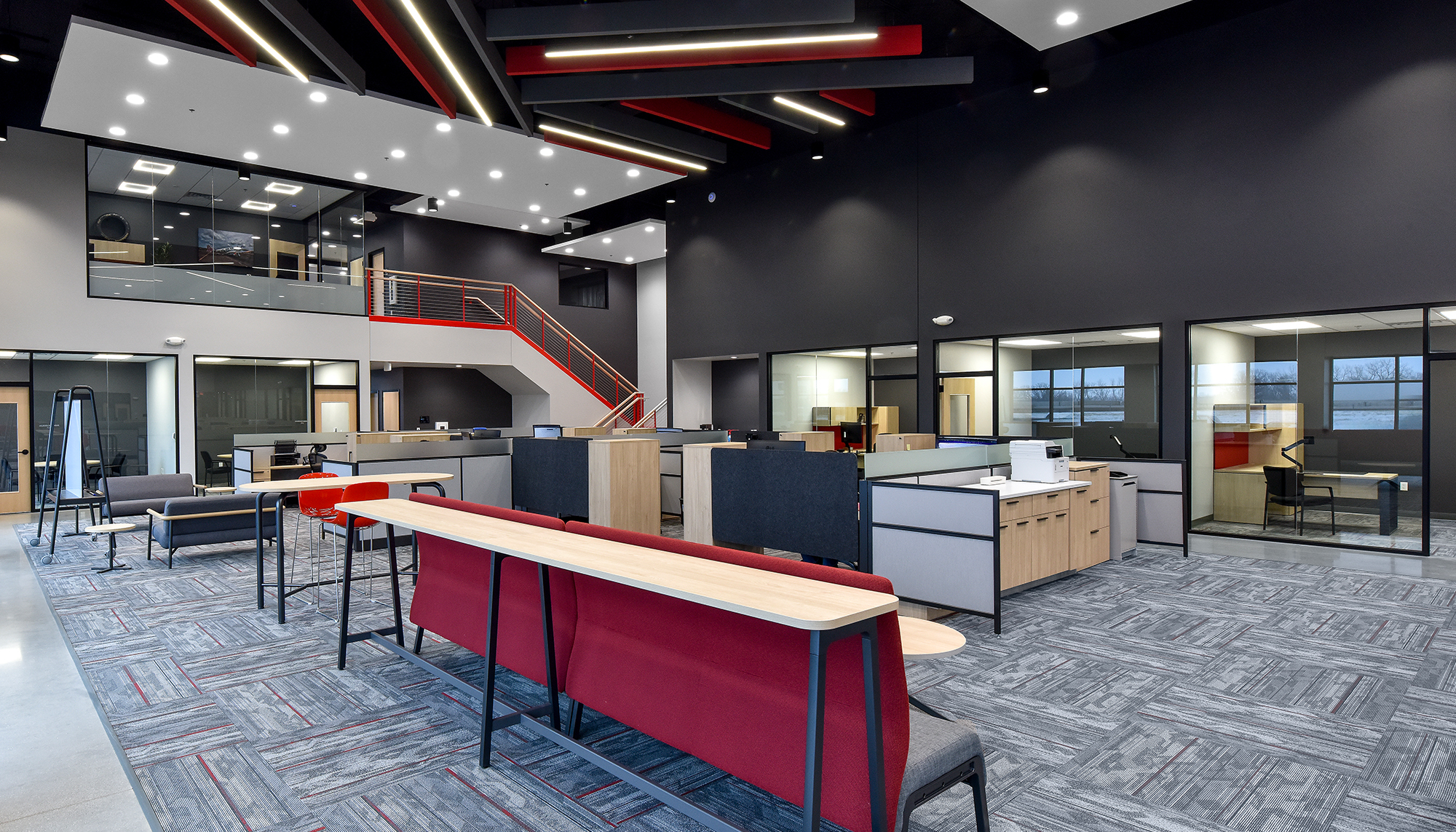 Modern open office with gray patterned carpet, black walls, red accents, shared workstations and a red staircase beneath geometric ceiling lights.