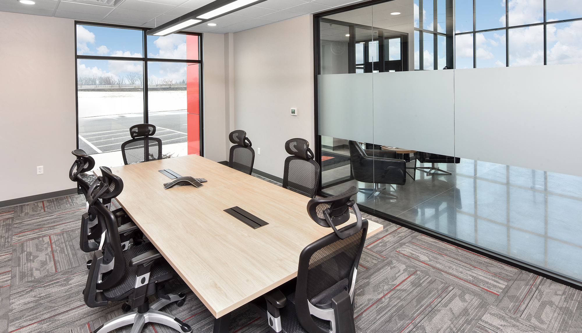 Modern conference room with a light wood table, black office chairs, gray patterned carpet and glass walls overlooking a bright atrium.