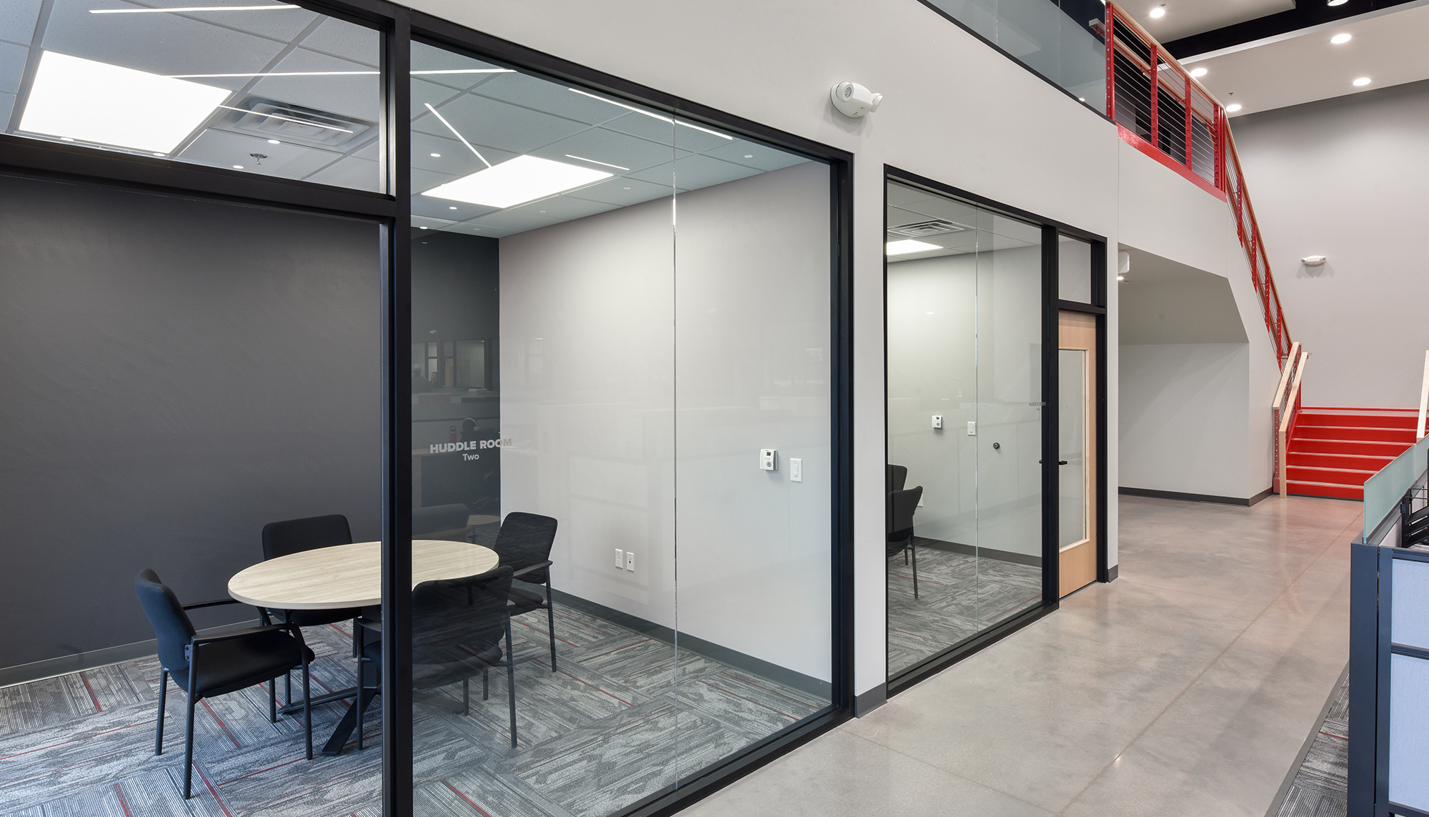 Glass-enclosed meeting room with black framing, gray carpet and a round table beside an open corridor with red stairs and railings.