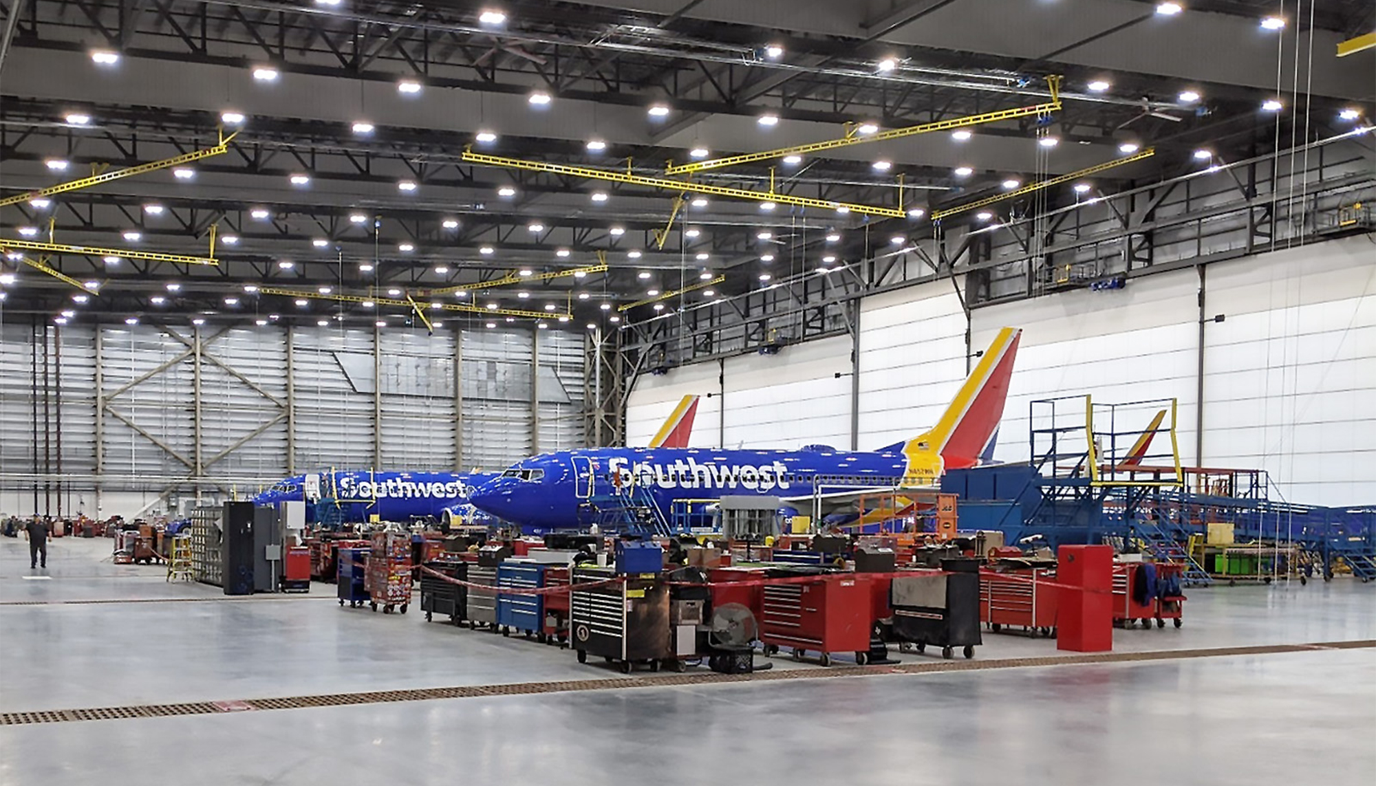 Large aircraft maintenance hangar with bright overhead lighting, yellow utility lines and a Southwest Airlines plane surrounded by maintenance equipment.