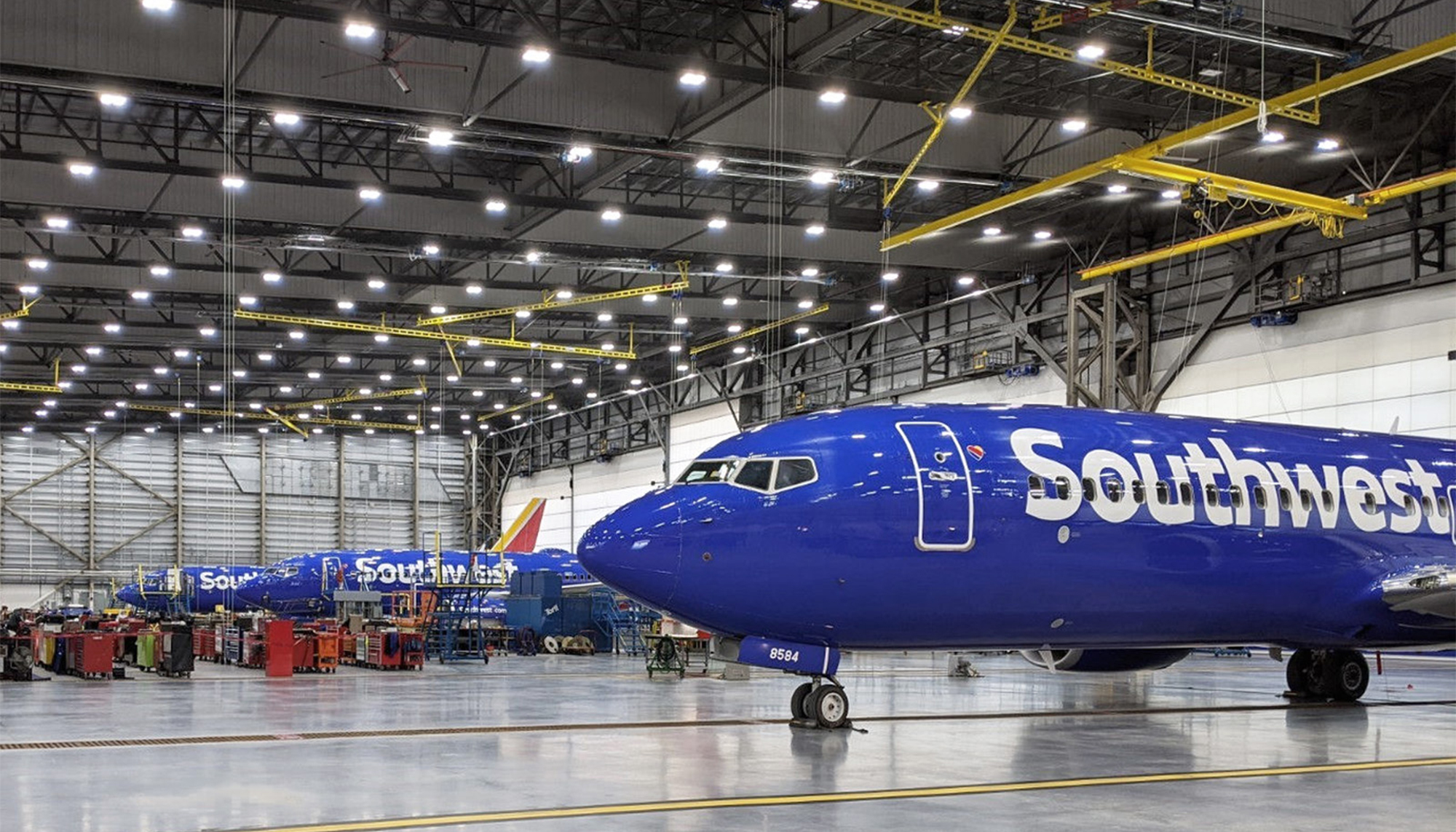 Blue Southwest Airlines plane inside a large maintenance hangar with black steel framing, yellow utility lines and bright overhead lighting.