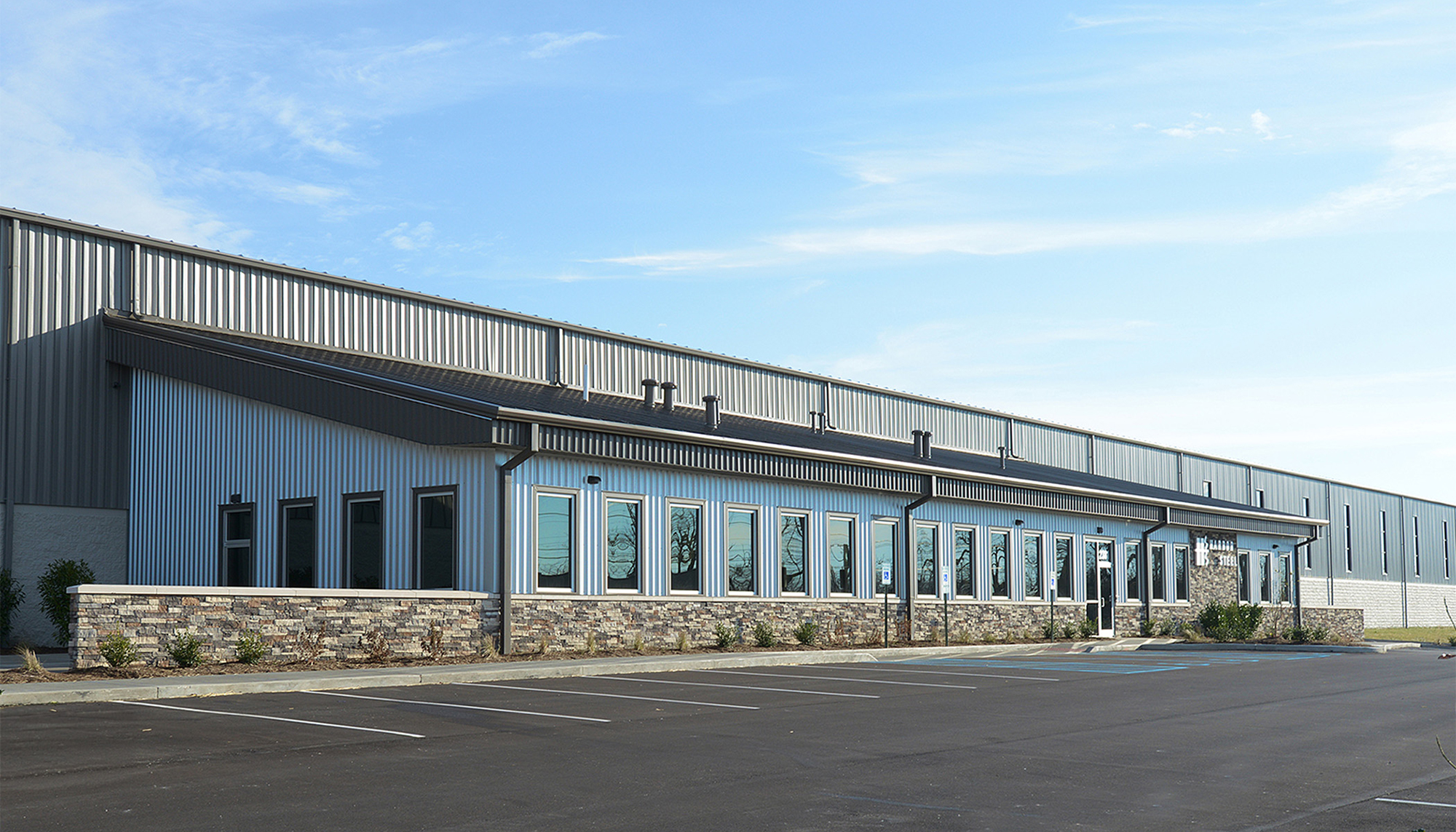 Exterior view of the Harbor Steel facility featuring metal wall panels, continuous storefront windows and stone facade accents.