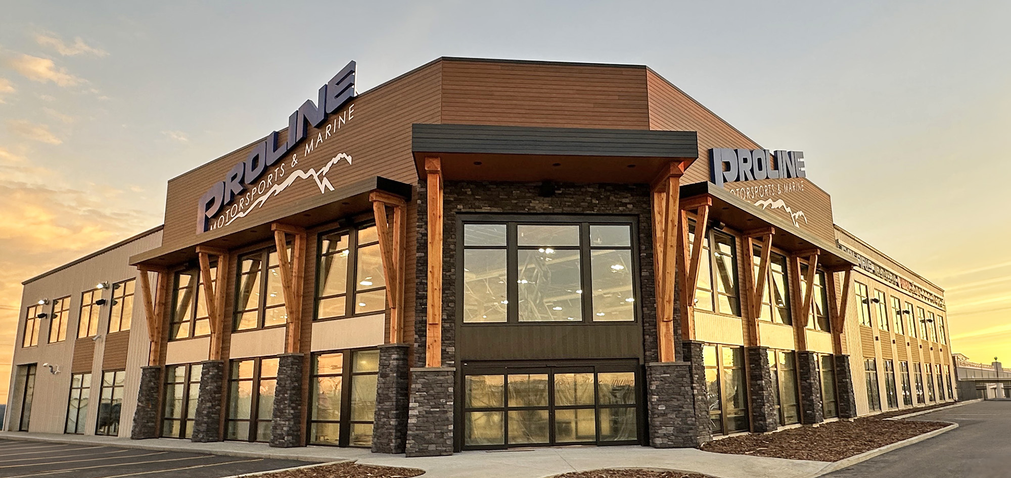 Proline Motorsports & Marine building featuring expansive glass windows, wood timber accents, stone columns and metal wall panels beneath a sunset sky.
