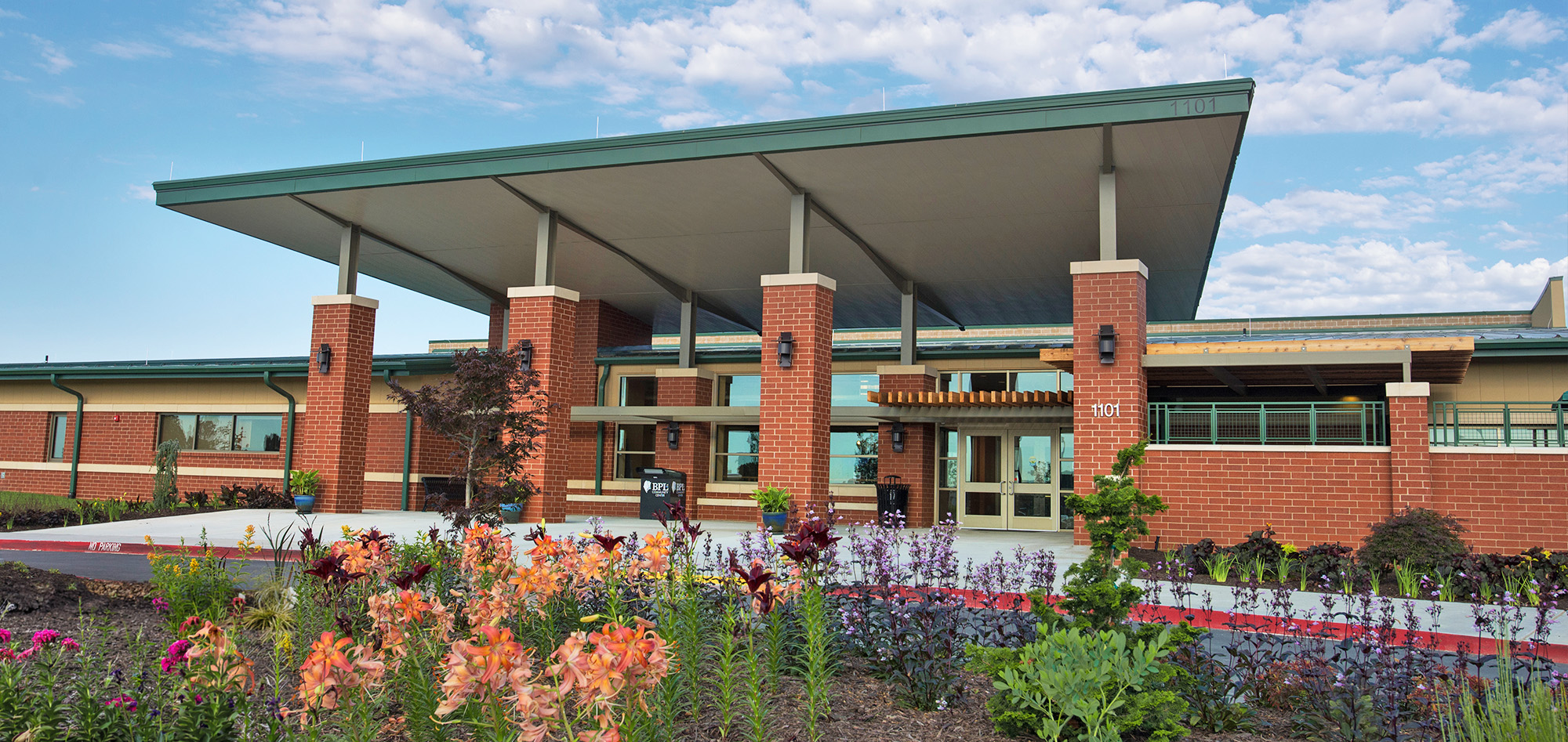 Red brick building entrance with a broad green metal roof, landscaped gardens and a blue, partly cloudy sky.